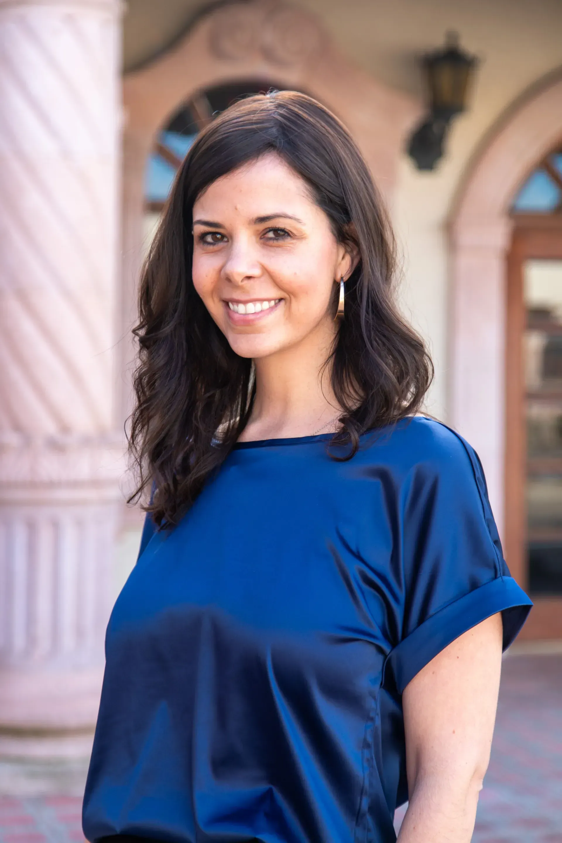 A woman in a blue shirt is smiling and standing in front of a building.