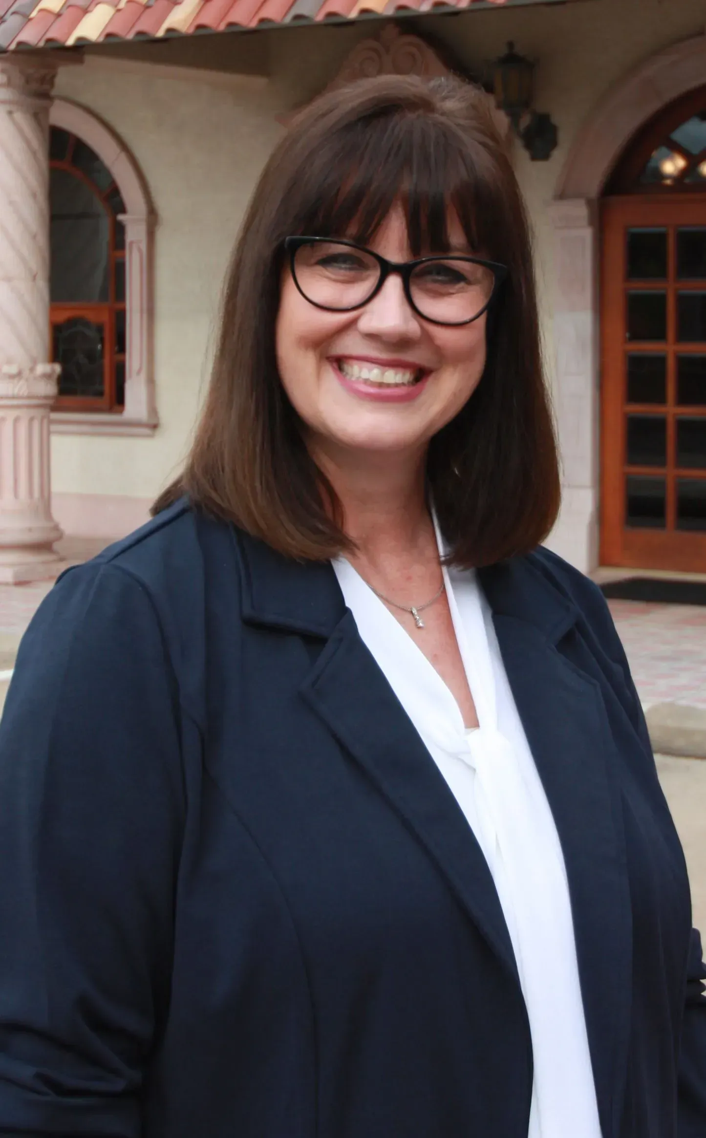 A woman wearing glasses and a blue jacket is smiling in front of a building.