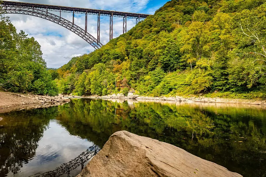 Wooded river gorge under a steel arch bridge, with calm water reflecting the landscape.