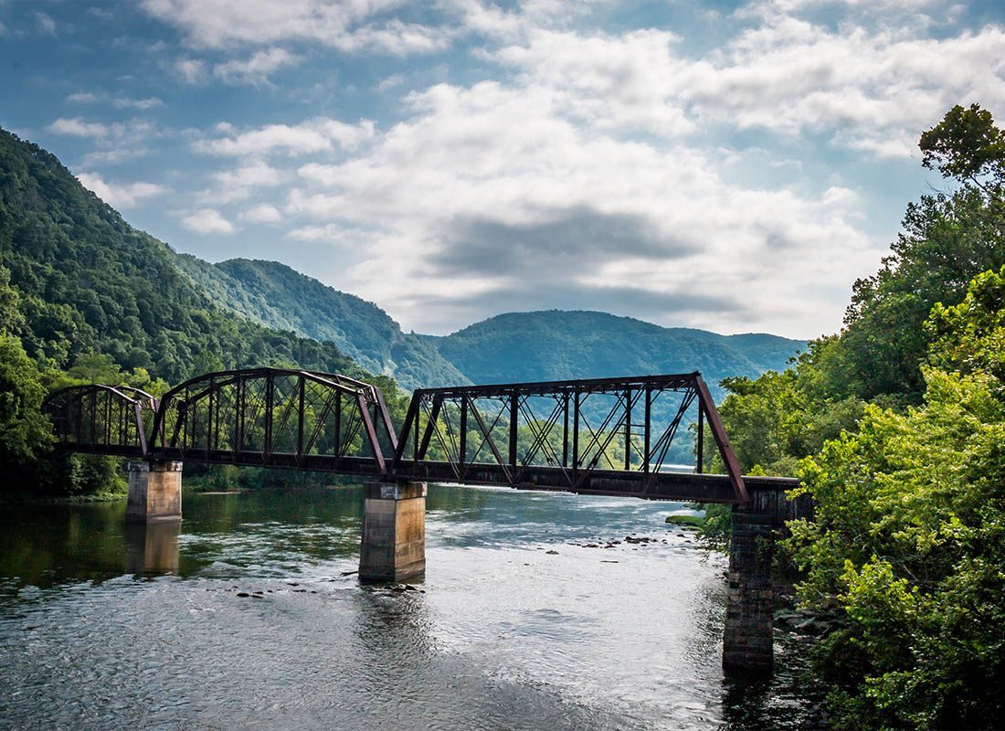 A steel bridge spans a river in a lush mountain valley under cloudy skies.