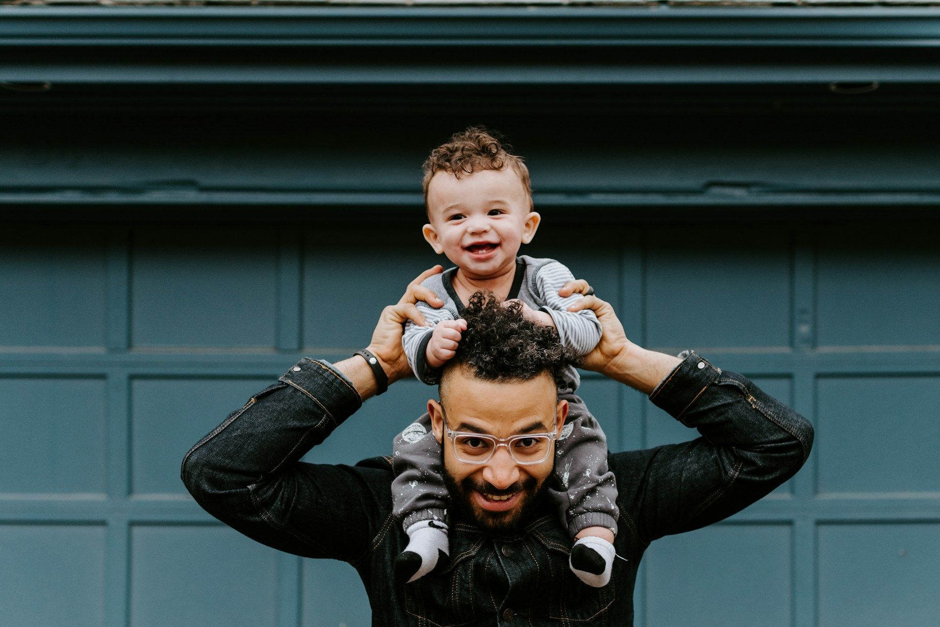 Man holding a smiling child on his shoulders in front of a blue garage door