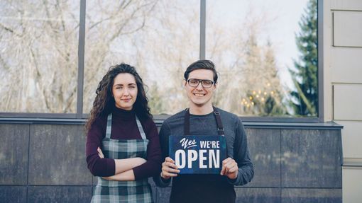 Two people standing outdoors, one holding a “WE’RE OPEN” sign.
