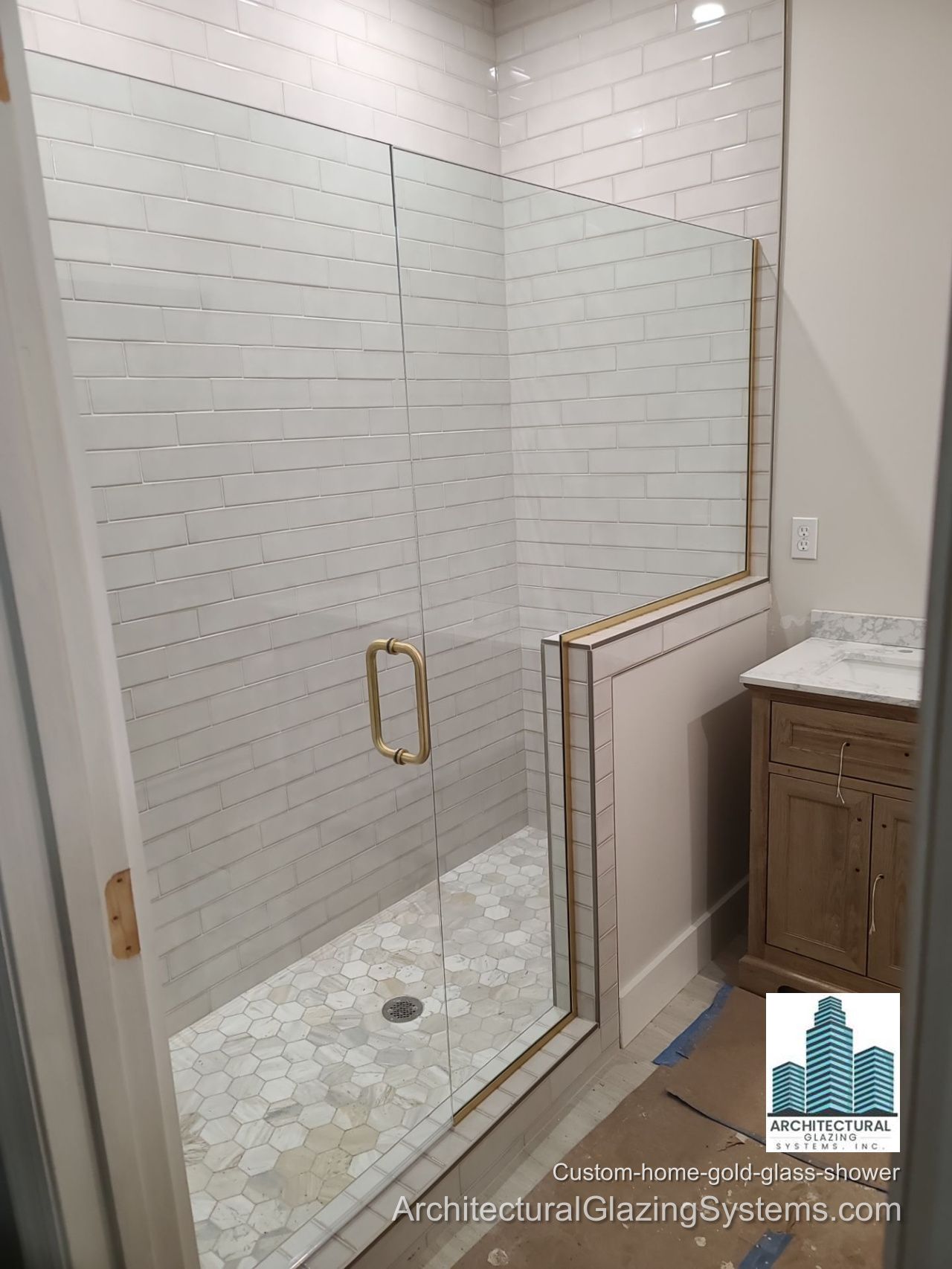Clear-glass shower enclosure with a gold-colored handle and trim beside white subway tile and a wood vanity.