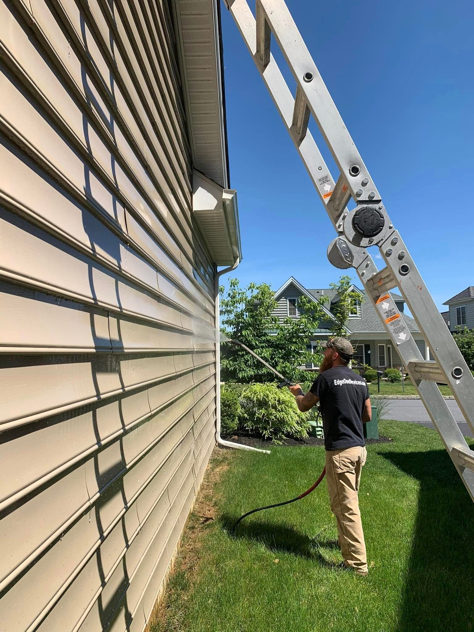 Person spraying a house siding with a hose while standing on the ground. A ladder is leaning against the house.