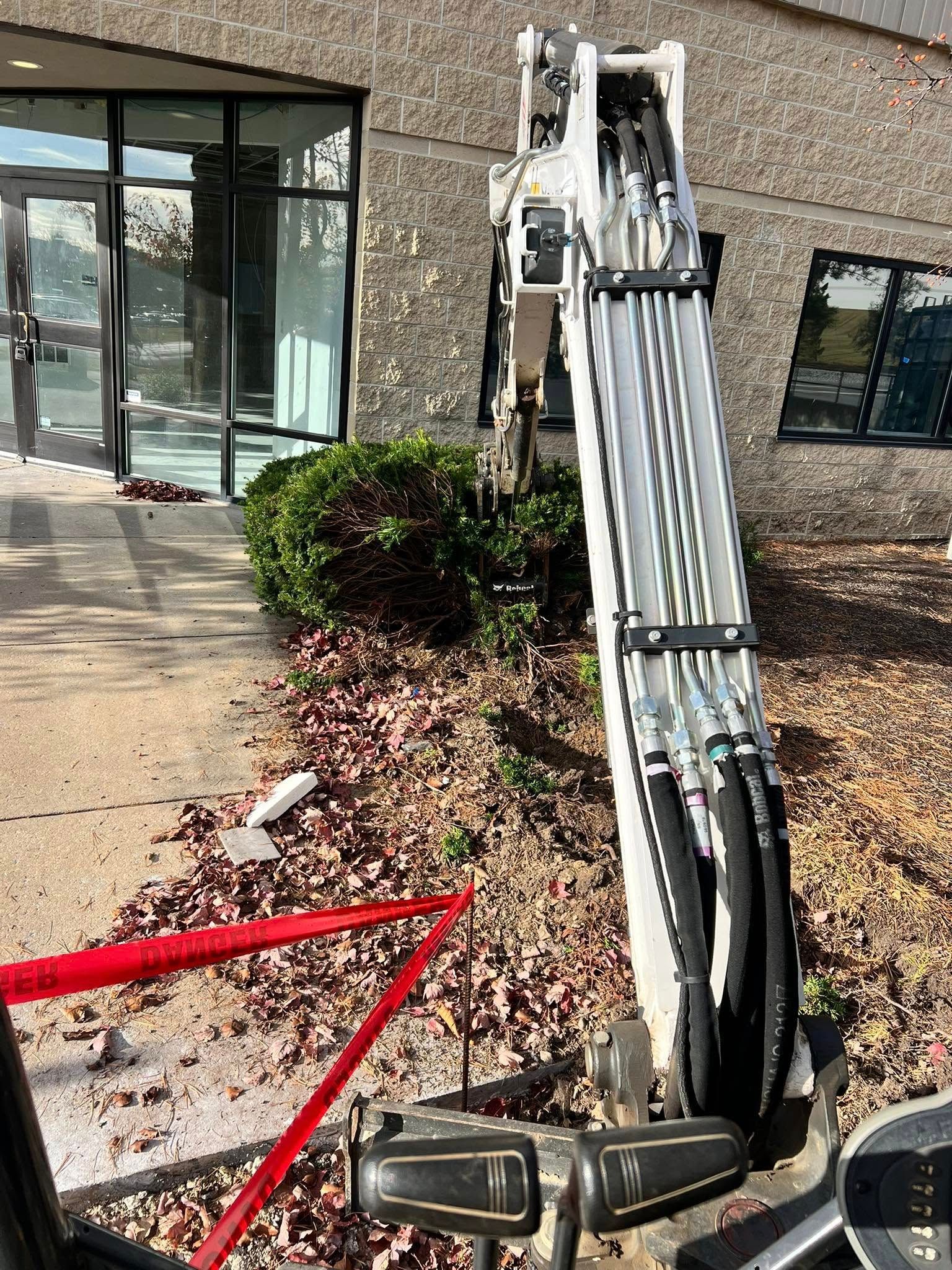 An excavator removing debris and a bush near a building entrance with glass doors.