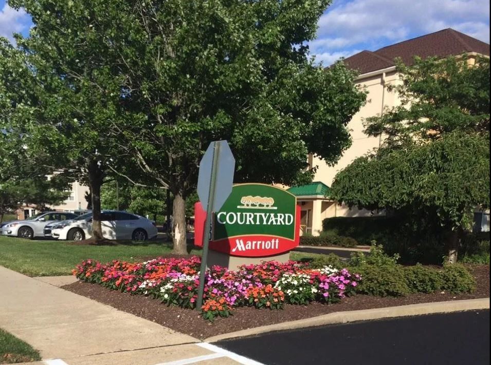 Courtyard by Marriott hotel sign with a floral border, under trees, with cars parked nearby.