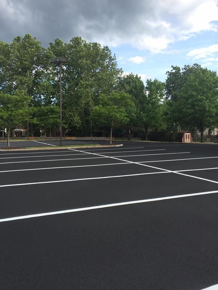 Empty asphalt parking lot with white painted lines; trees and cloudy sky in background.
