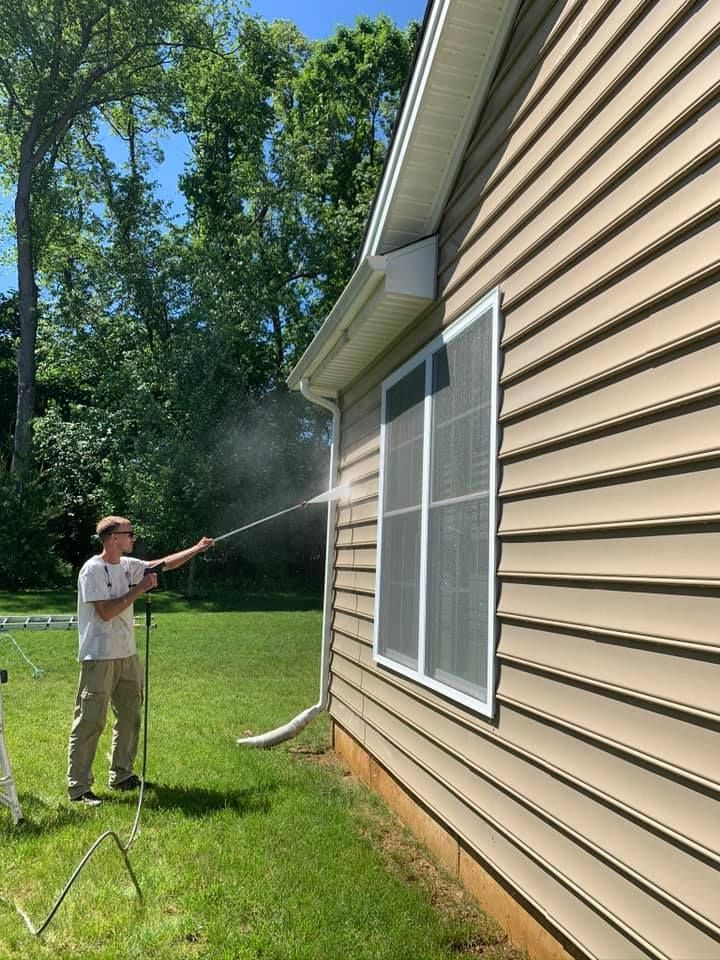 Man power washing the side of a beige house with a window, spraying the gutters. Outdoors.