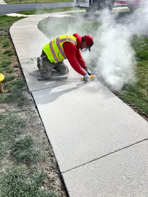 Worker in safety vest and mask cutting concrete sidewalk, creating dust.