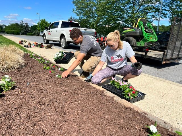 Two people planting flowers in a brown mulched garden bed. White truck, mower trailer in background.