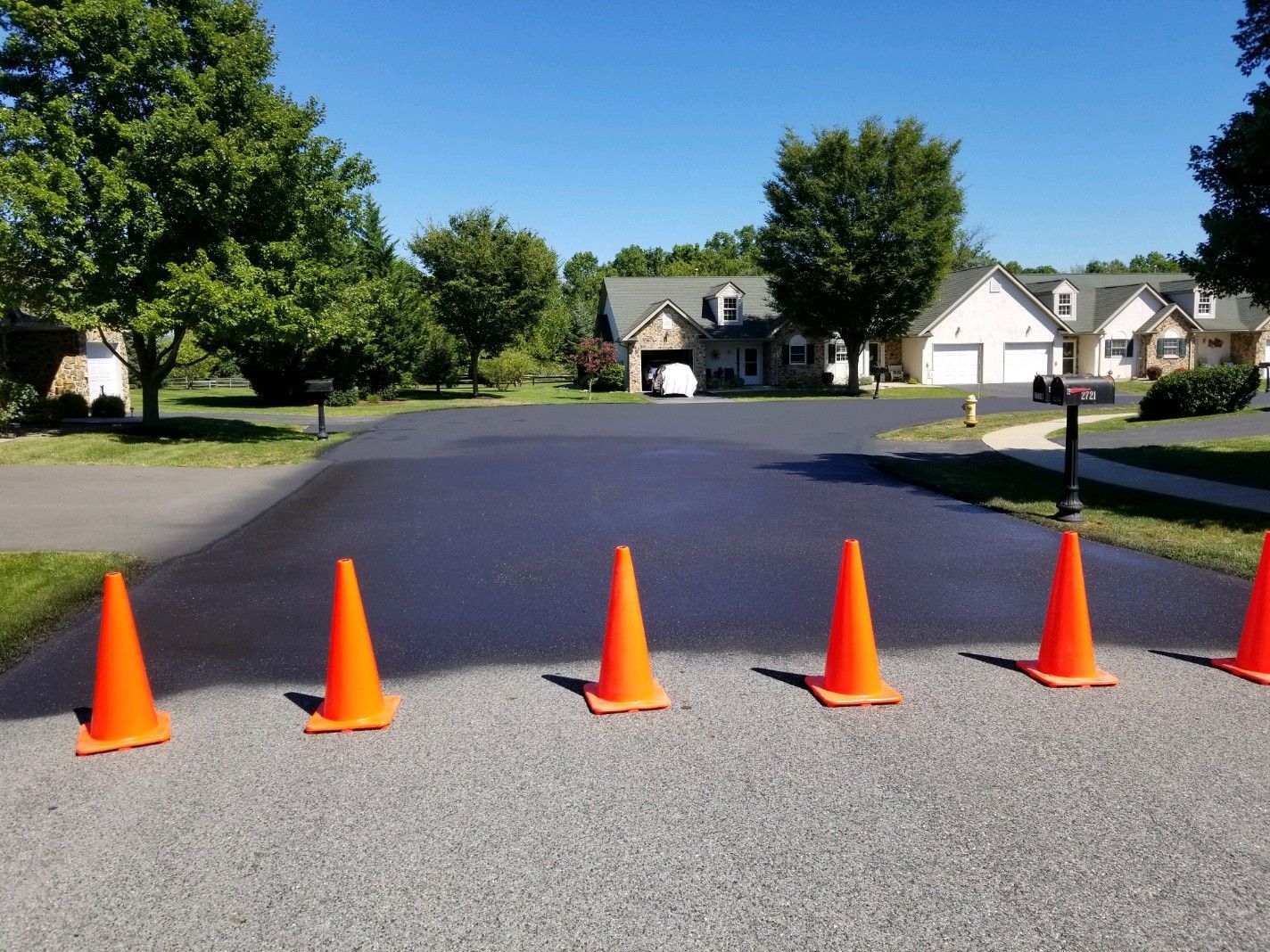 Newly paved asphalt driveway with orange traffic cones. Houses and trees in the background.