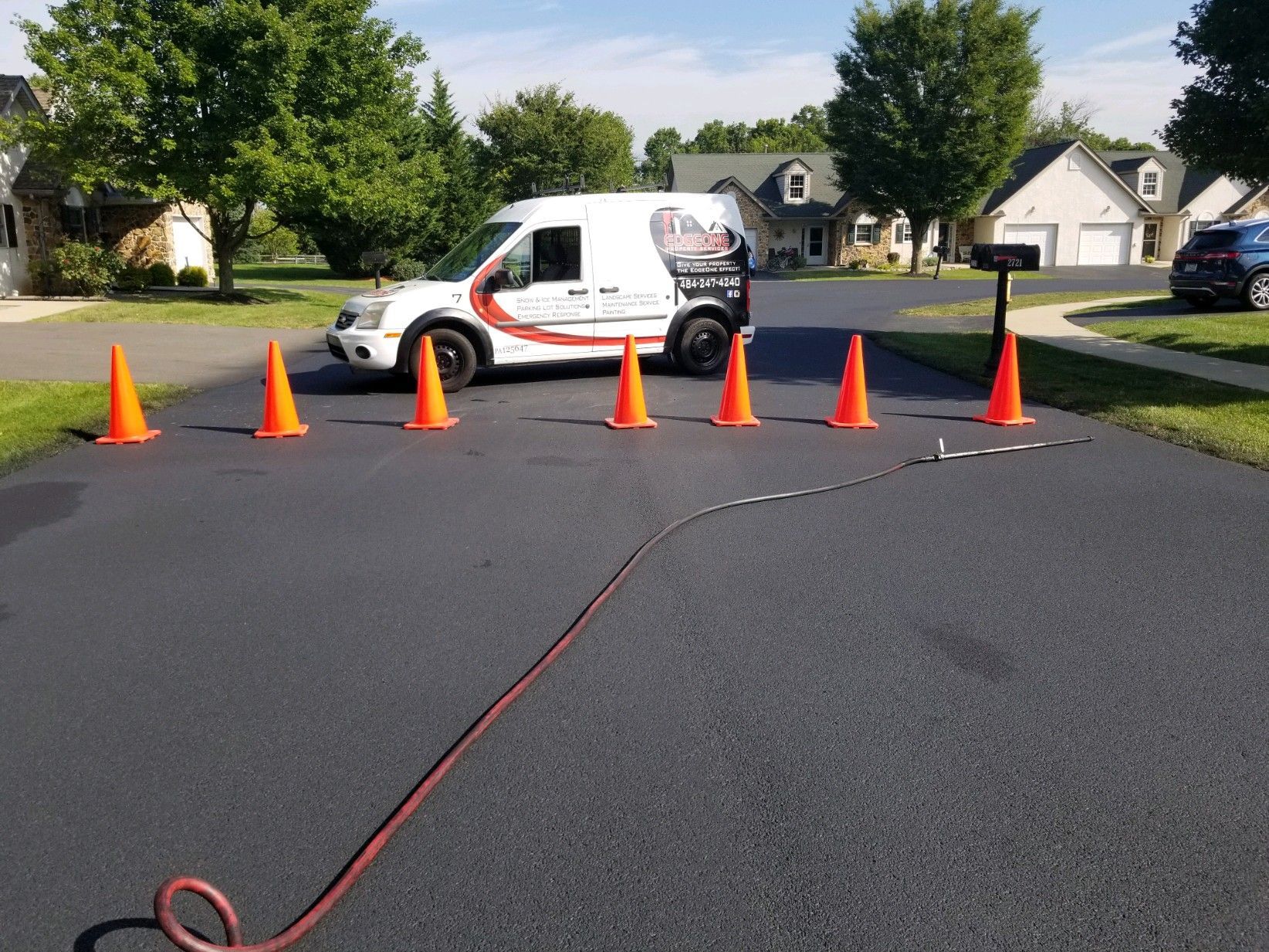 A white service van parked on a driveway with orange cones and a hose, suburban setting.