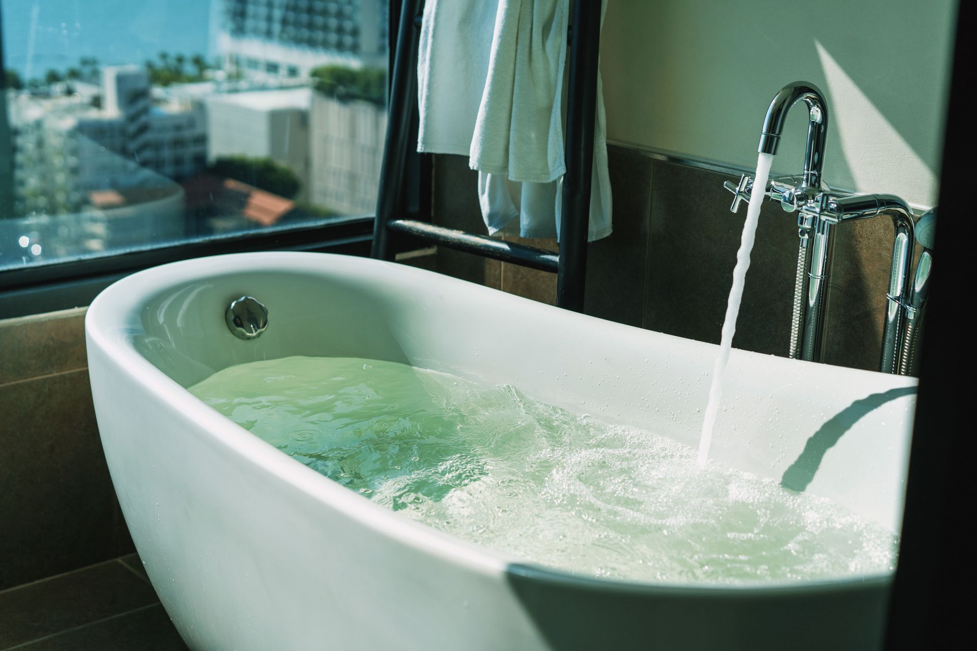 A bathtub, by a panoramic window overlooking the city skyline, is being filled with water.