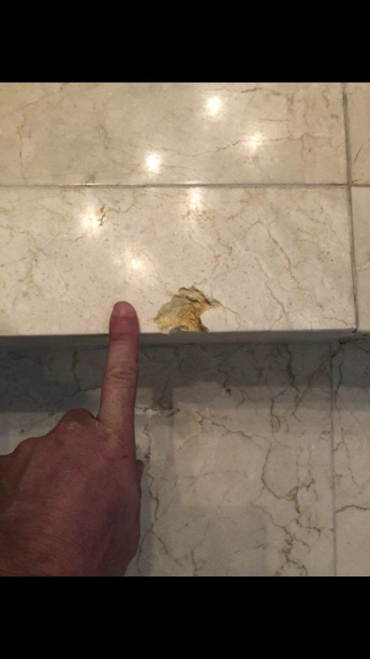 Finger pointing at a small yellow leaf or debris on a beige tiled floor near a step.