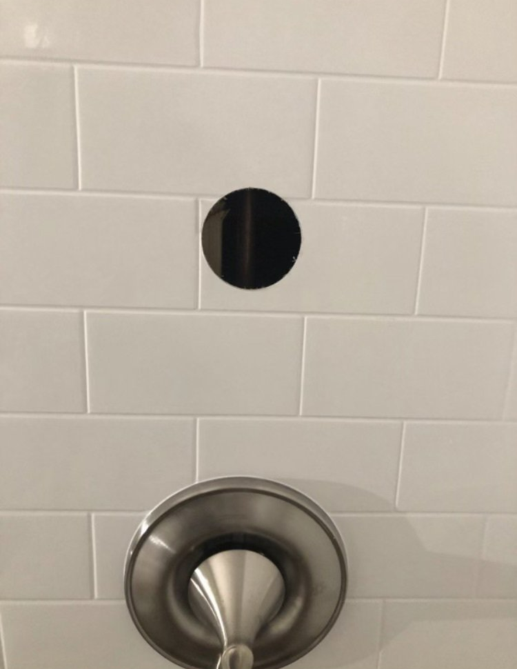 White tiled shower wall with a circular black opening above a chrome shower fixture.