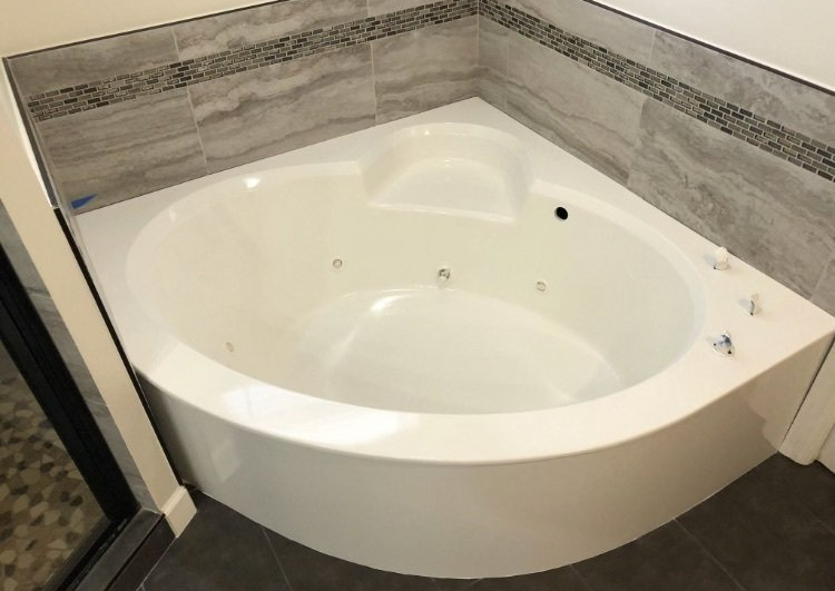 Corner white jetted bathtub in a tiled bathroom with gray walls and dark floor tile