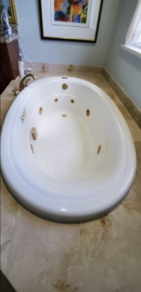 White oval bathtub with gold jets in a tiled bathroom corner near a window and framed art