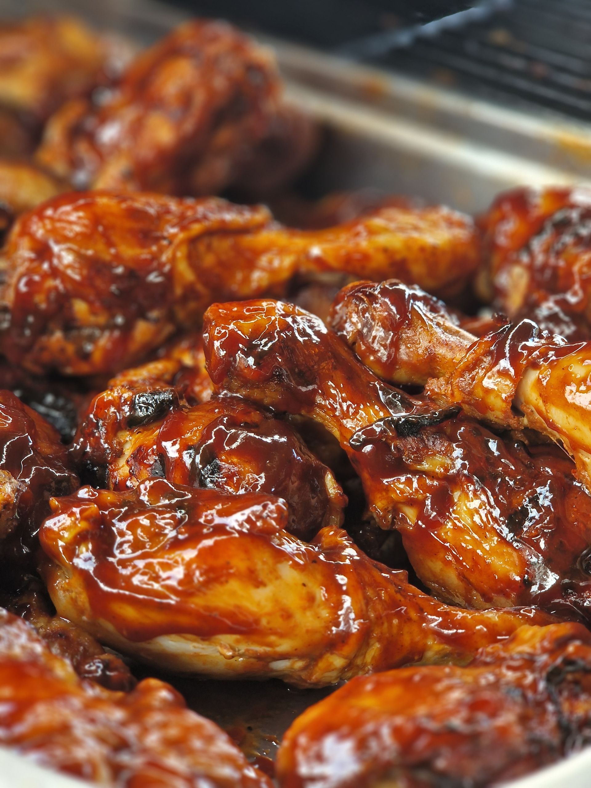 Een close-up van meerdere kippenpootjes, bedekt met een dikke, donkerbruine barbecuesaus, die in een metalen schaal liggen.