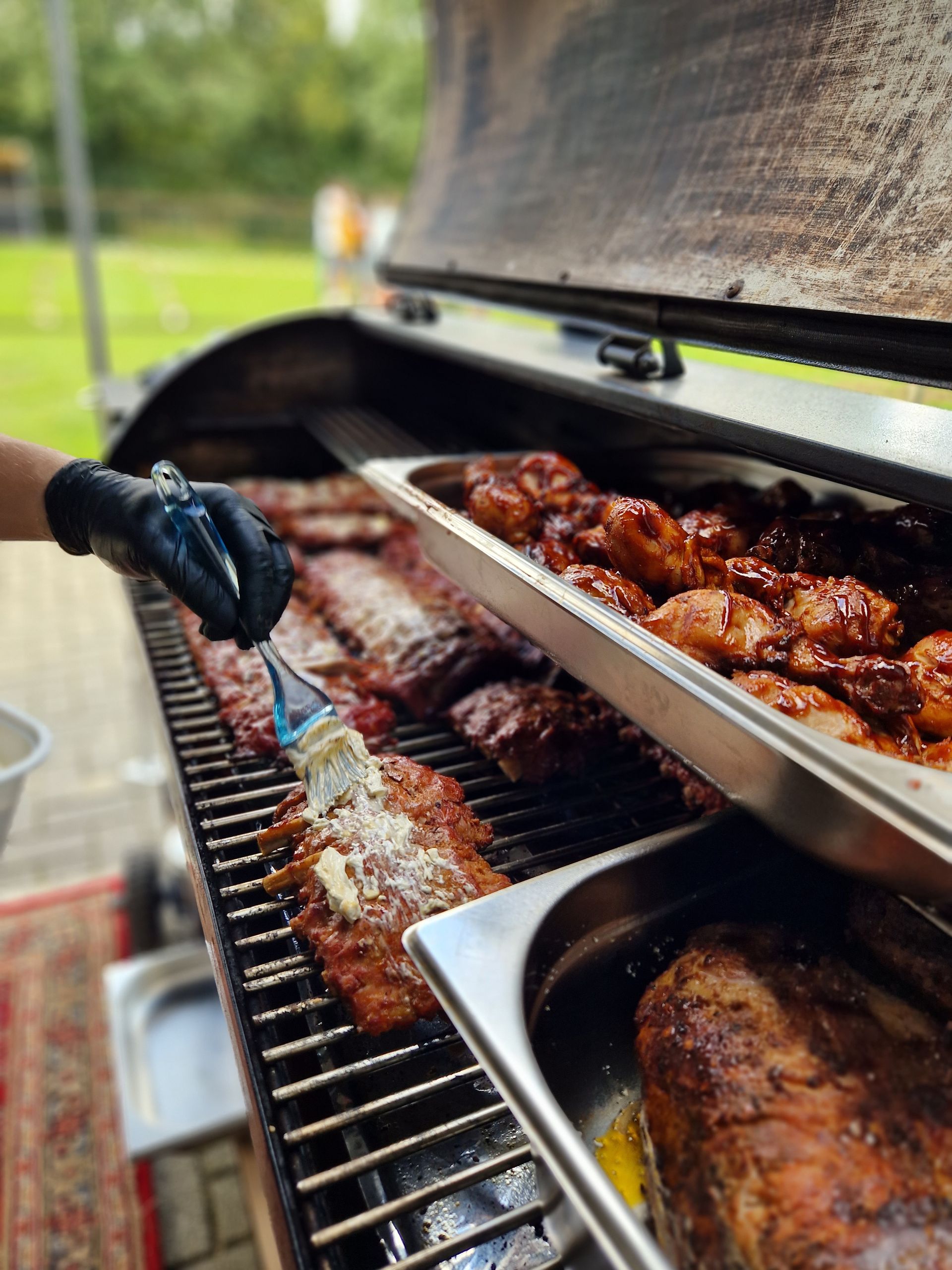 Een persoon met een zwarte handschoen gebruikt een kwast om ribben te bestrijken op een barbecue, waarboven een schaal met vlees aan het garen is.