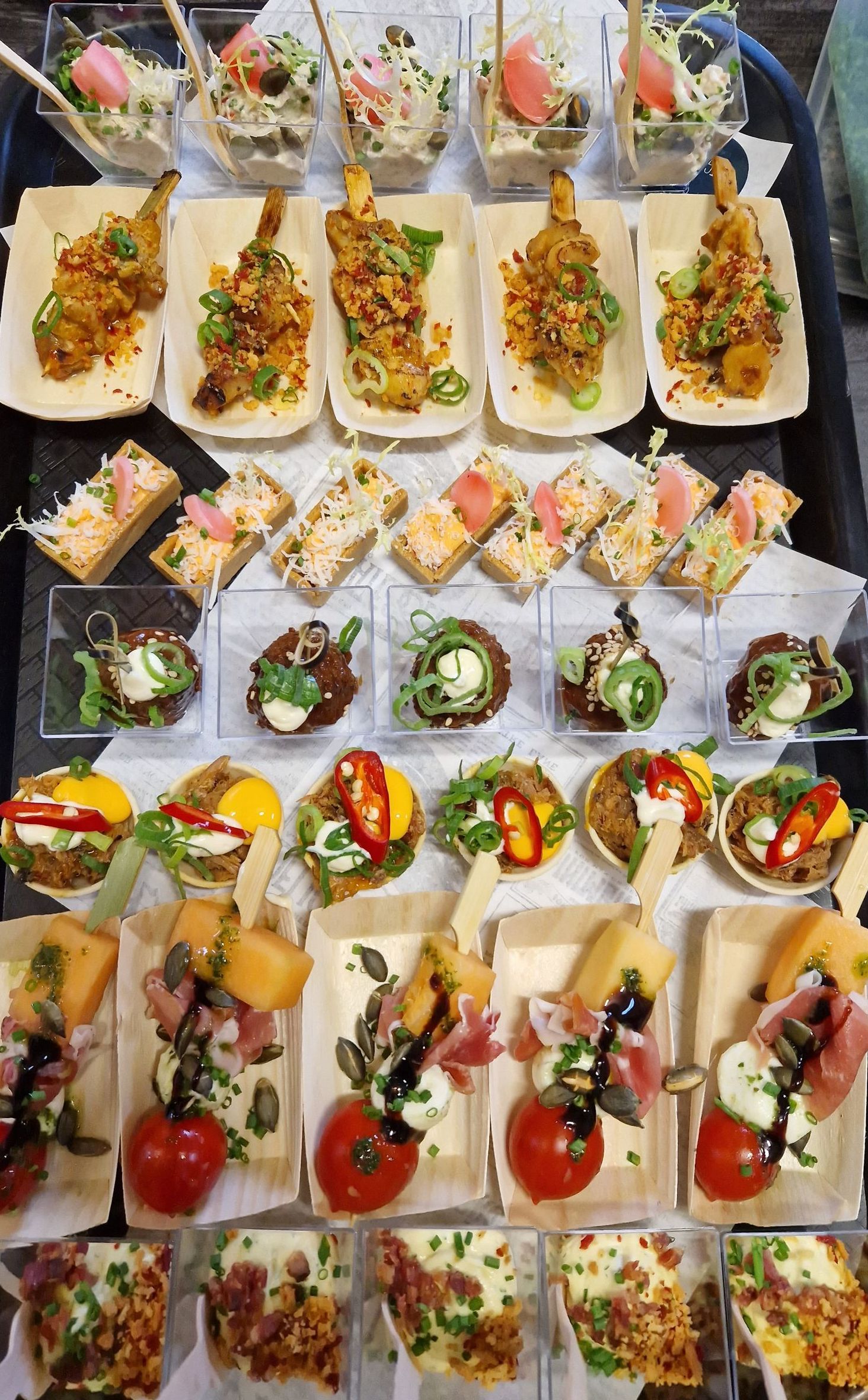 Een assortiment van gastronomische hors d'oeuvres, in rijen gerangschikt op een zwart dienblad, waaronder spiesjes, crostini en kleine glaasjes.