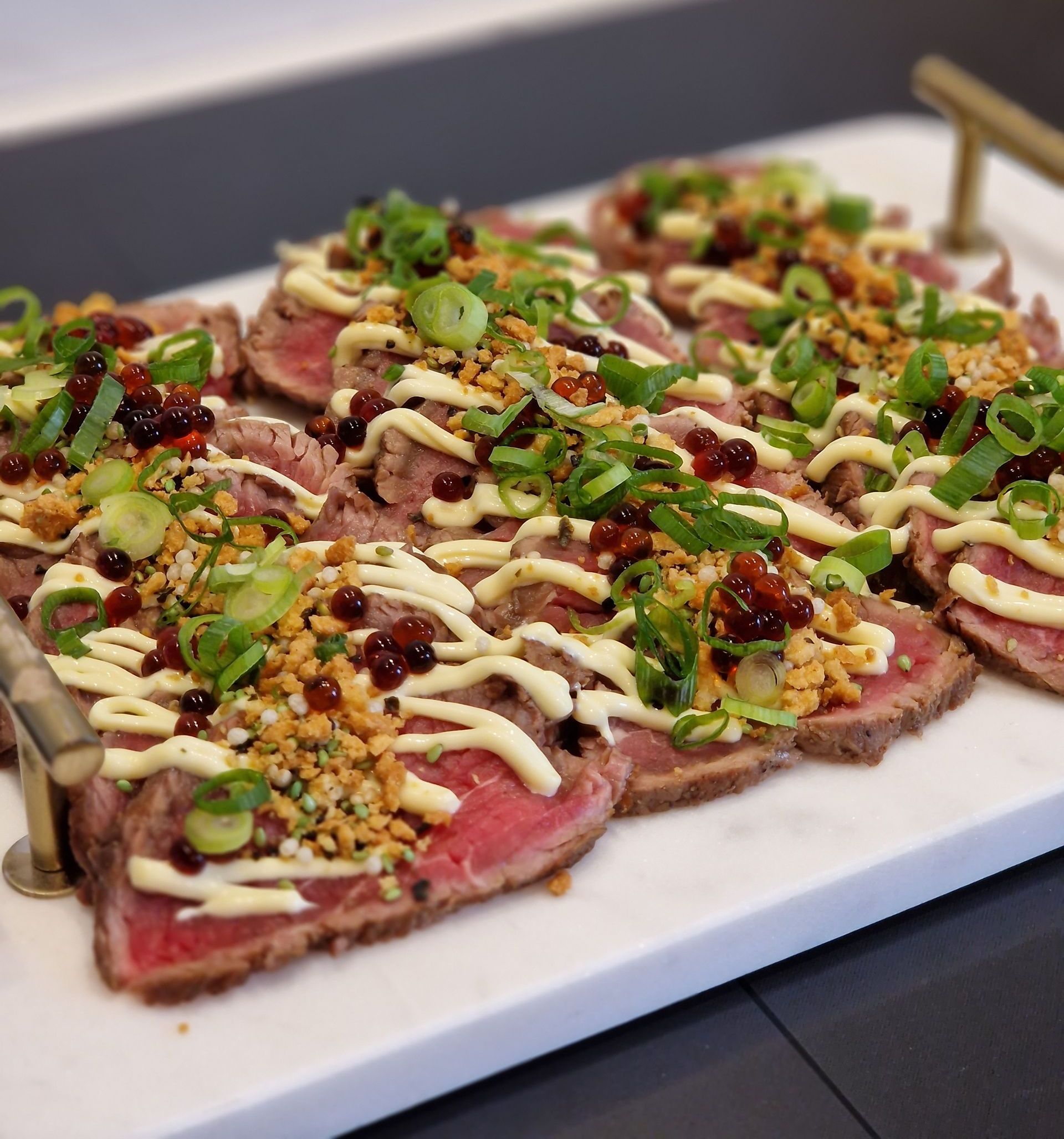 Dun gesneden tataki van rauw rundvlees, gegarneerd met lente-uitjes, krokante knoflook, een scheutje witte saus en donkere parels, geserveerd op een schaal.