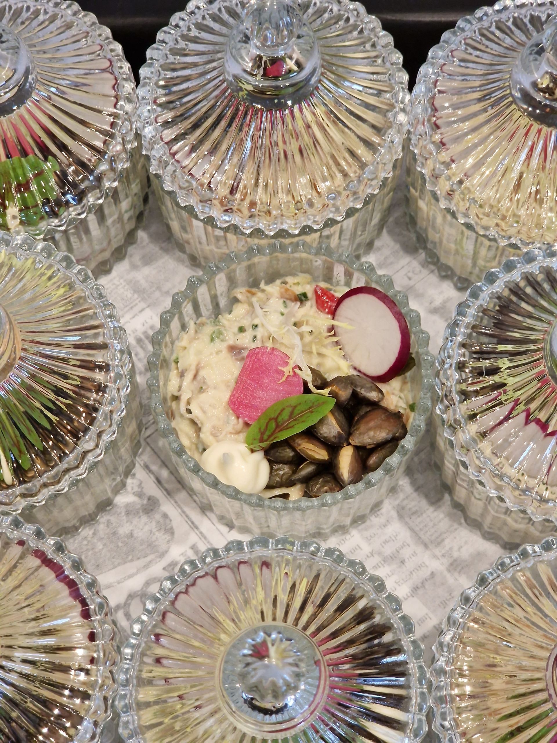 Een opname vanuit een hoog perspectief van meerdere kristallen schalen met deksels, waarbij één van de middelste schalen open is en een salade met radijs onthult.