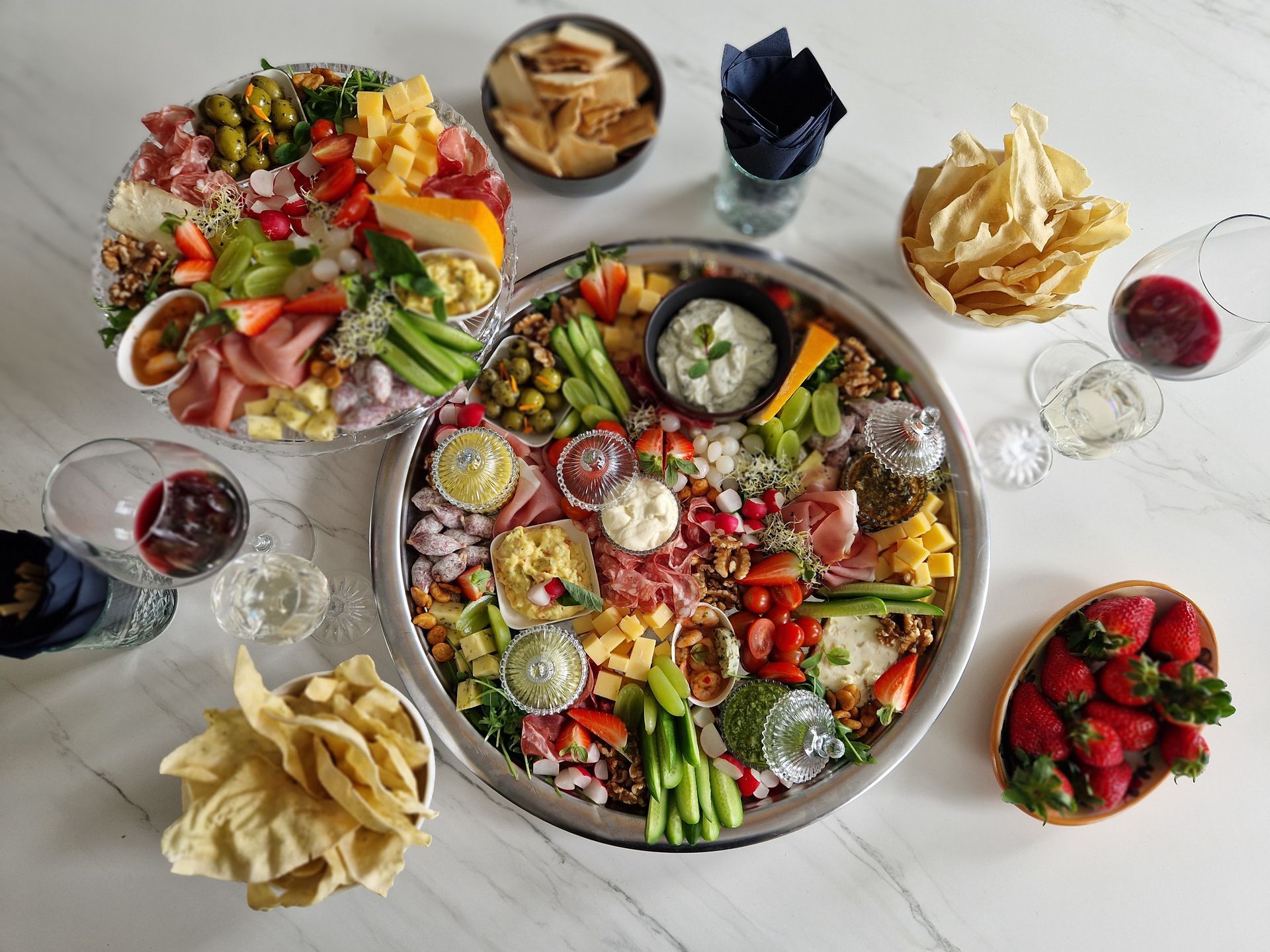 Twee charcuterieplanken met vleeswaren, kazen, groenten en dipsauzen, omringd door chips, aardbeien en glazen wijn.
