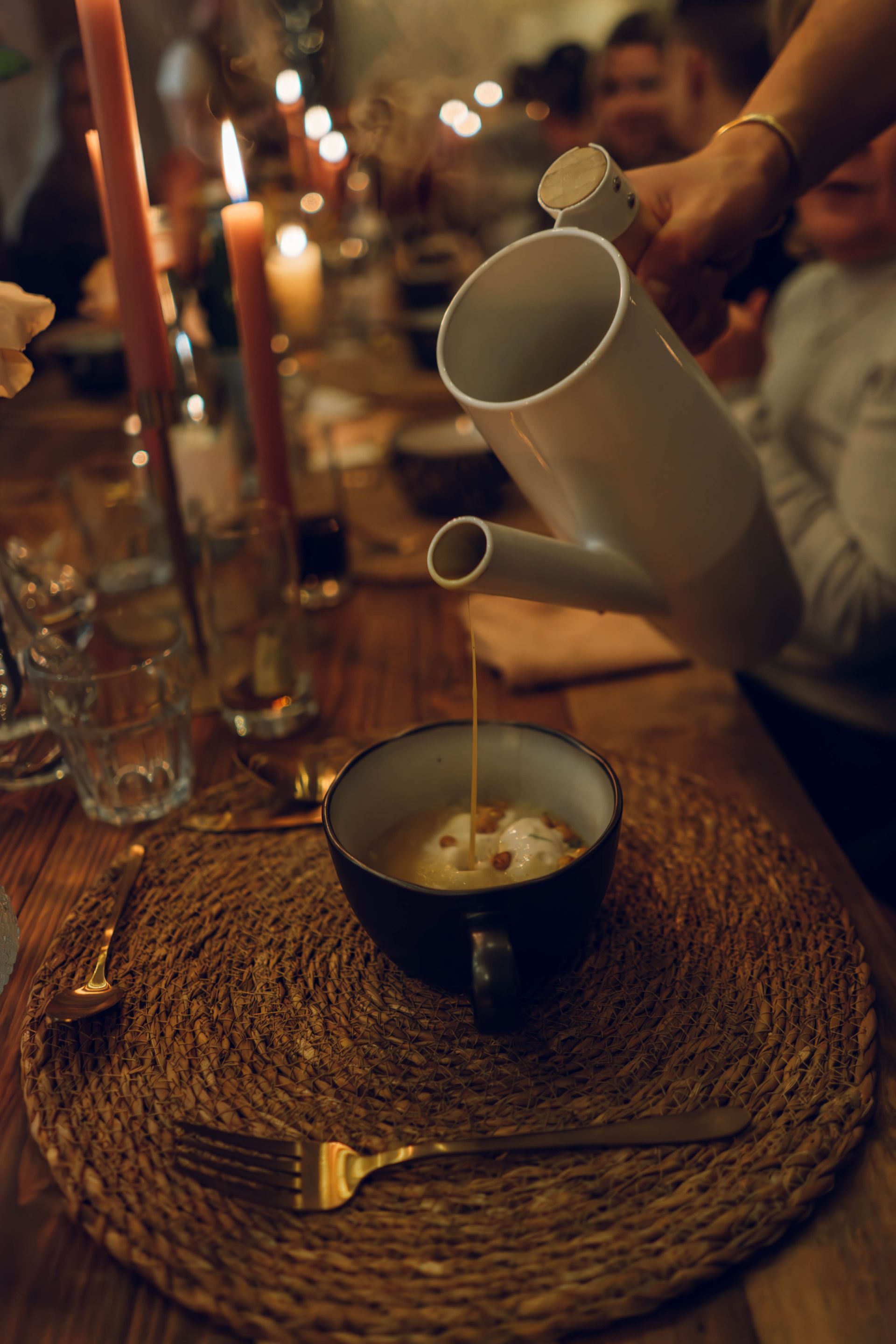 Een hand schenkt vloeistof uit een witte keramische kan in een donkere kom op een geweven placemat aan een eettafel bij kaarslicht.