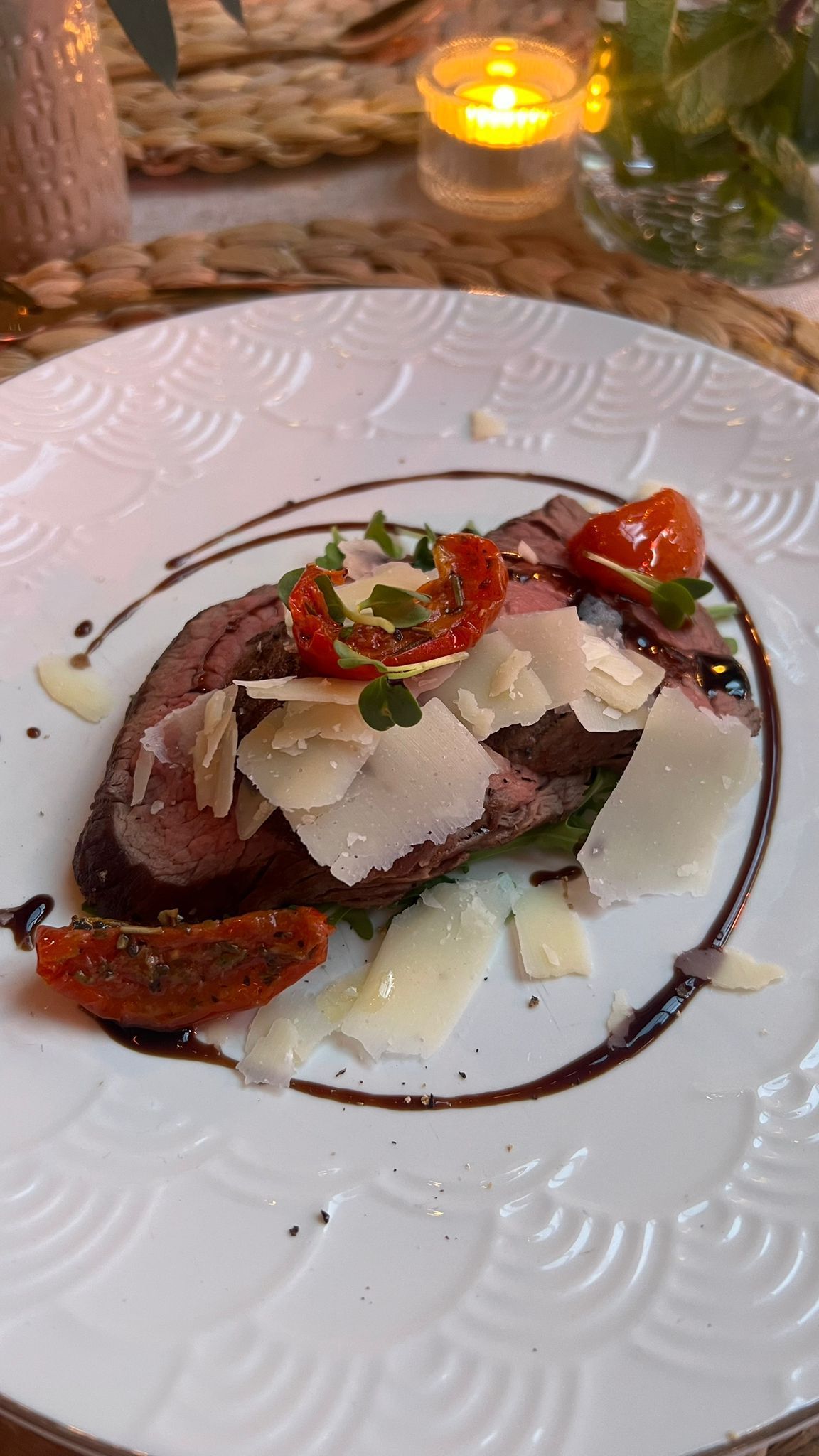 Gesneden biefstuk, gegarneerd met geraspte Parmezaanse kaas en zongedroogde tomaten, geserveerd op een wit bord met reliëf en besprenkeld met balsamico glazuur.