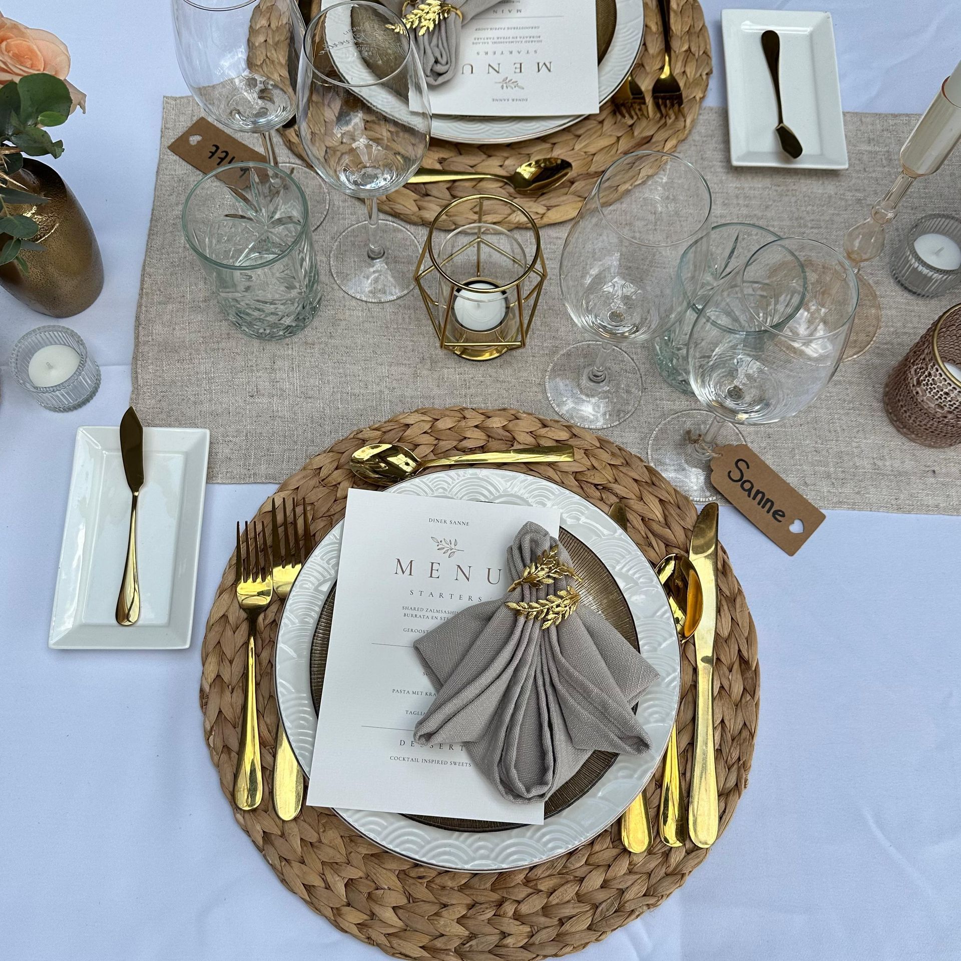 Een elegante, formele tafelsetting met geweven placemats, witte borden, gouden bestek, glazen en gedrukte menu's.