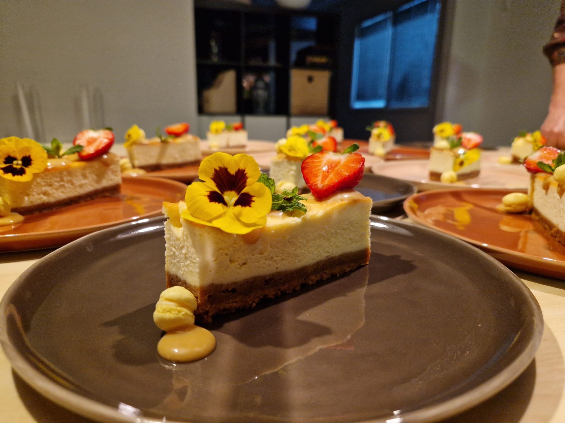 Plakjes cheesecake, gegarneerd met gele viooltjes en aardbeien, met een toefje slagroom op een donker bord.