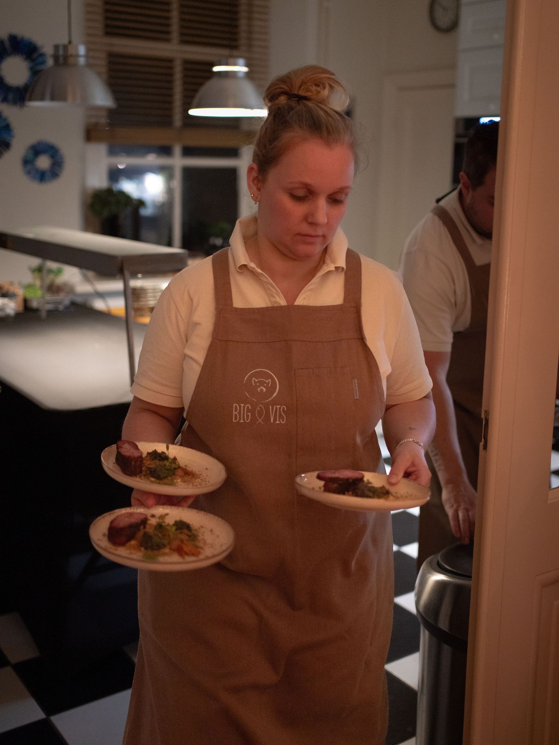 Een ober met een beige schort loopt over een geblokte vloer en draagt ​​drie borden met eten.