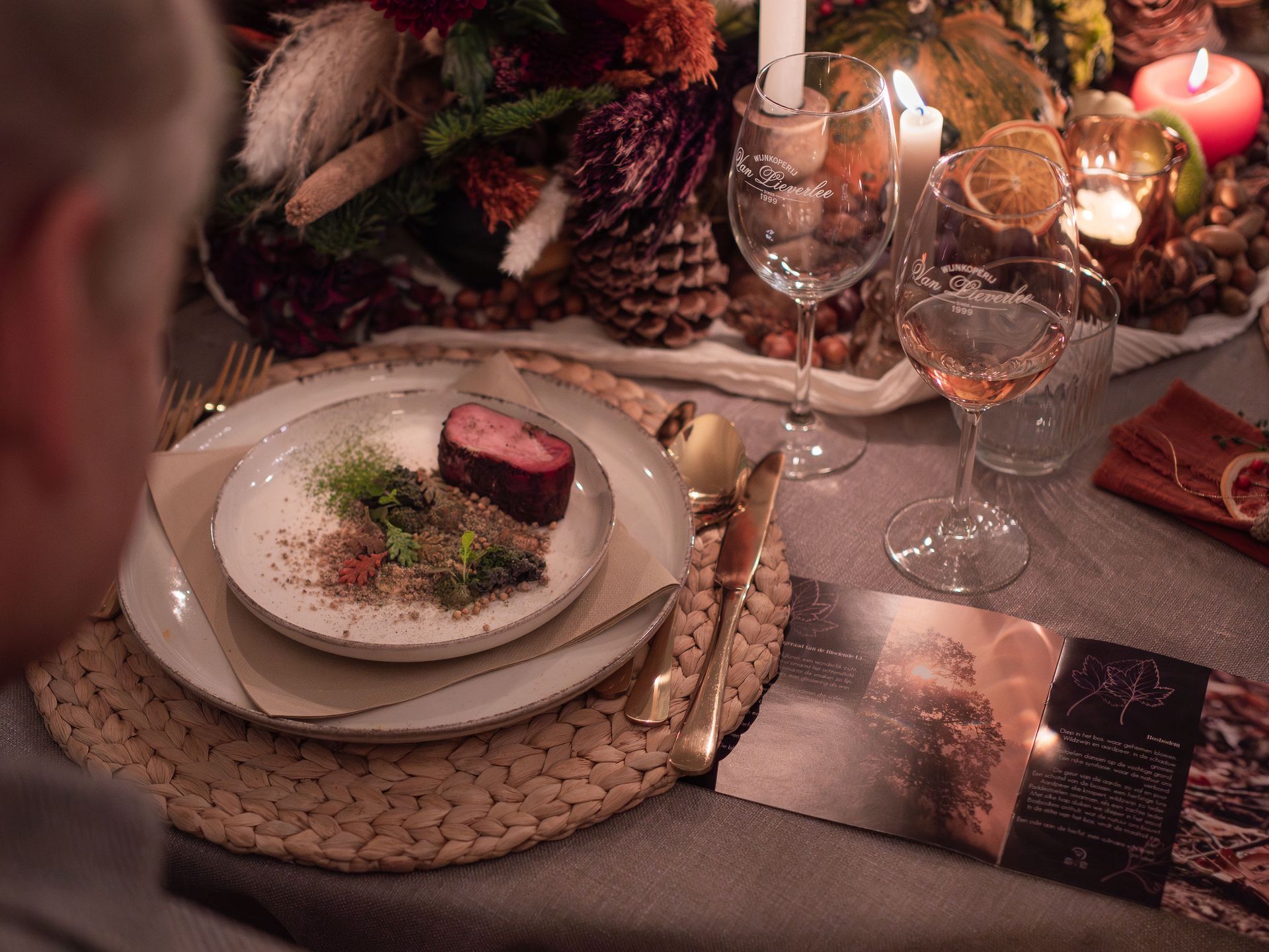 Iemand zit aan een tafel die bij kaarslicht is gedekt voor het diner, met een opgediende maaltijd, wijn en een decoratief herfststuk als tafeldecoratie.