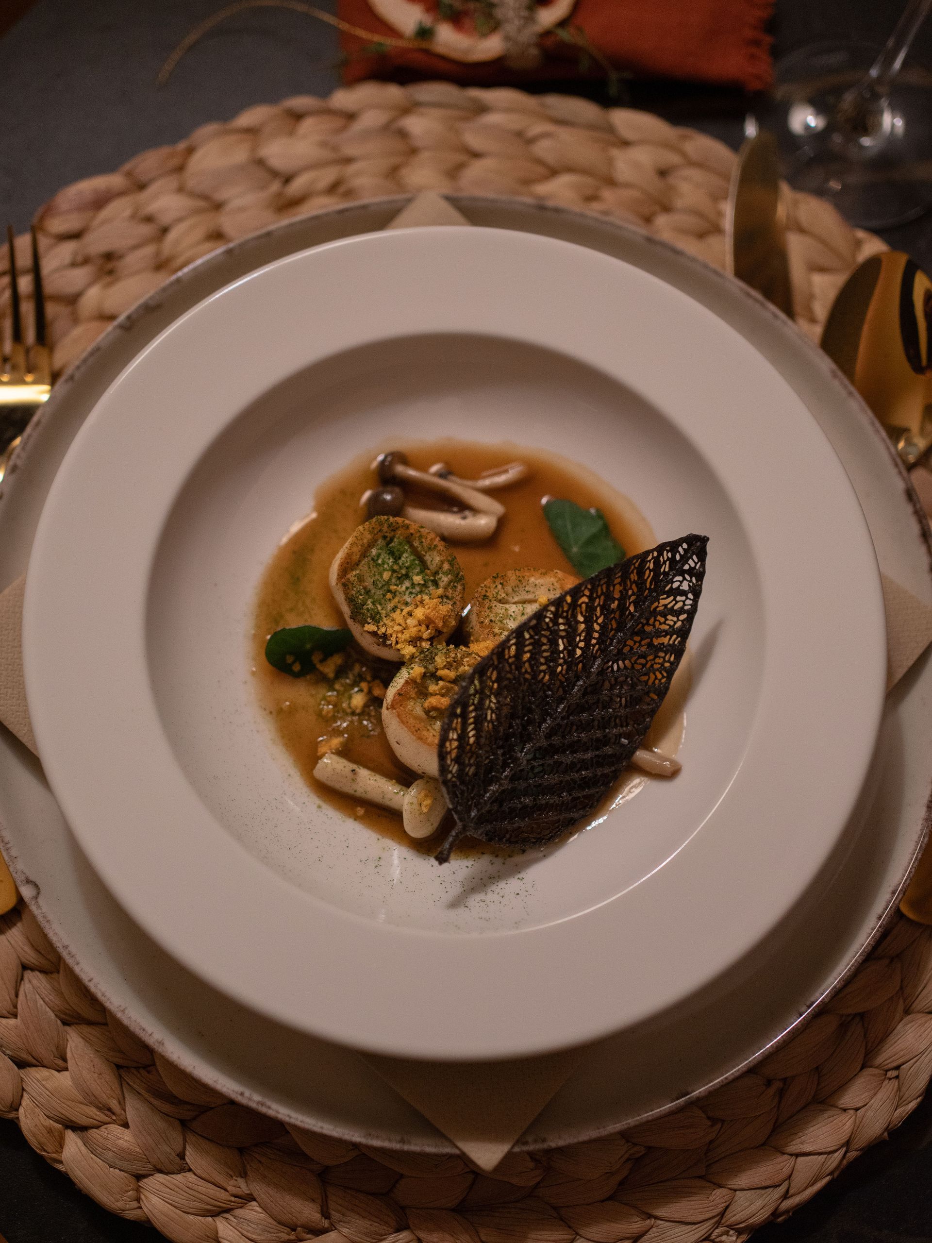 Een gastronomisch gerecht met gevulde pasta, champignons en een decoratieve donkere kanten tuile in een witte kom op een geweven placemat.