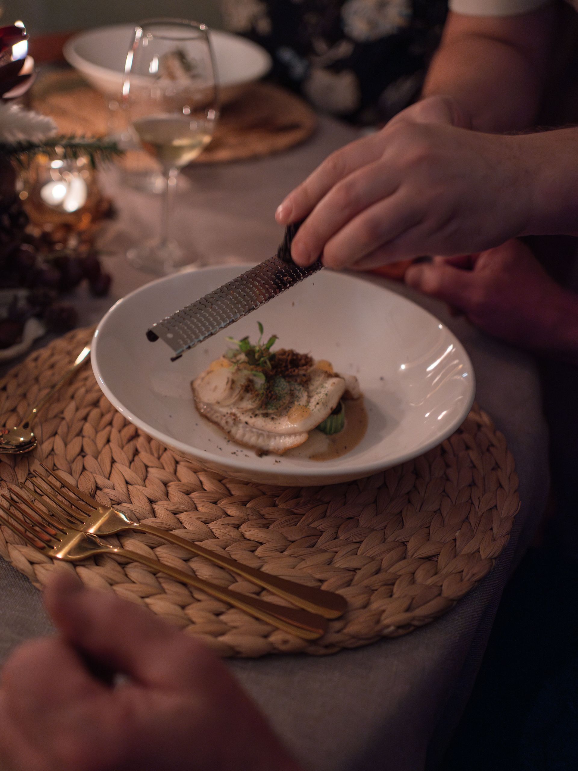 Een hand gebruikt een microplane om garnering over een stuk vis te raspen dat in een witte kom op een geweven placemat wordt geserveerd.