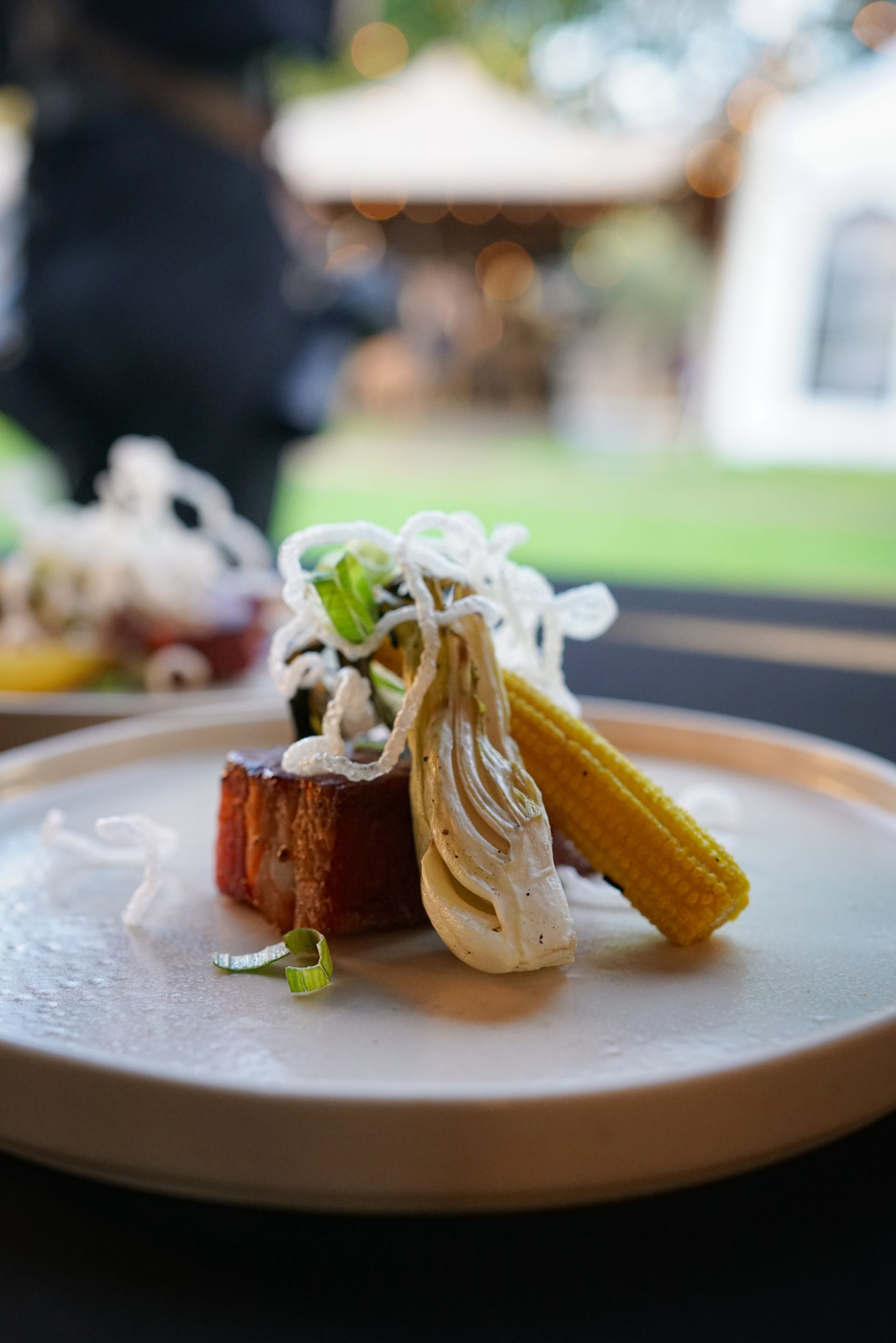 Een gastronomisch gerecht met een stuk buikspek, mini-maïs en garnering, geserveerd op een donkere tafel tijdens een evenement in de buitenlucht.