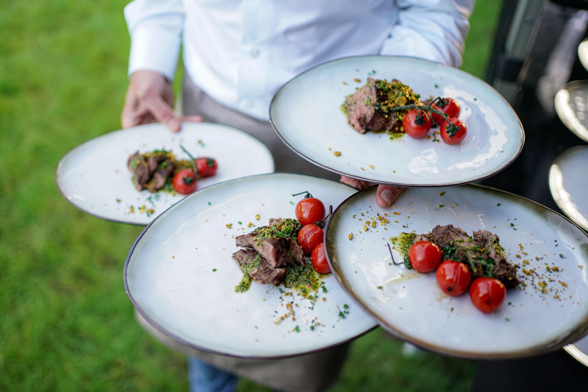 Een ober houdt drie borden met biefstuk en cherrytomaten vast op een grasveld.