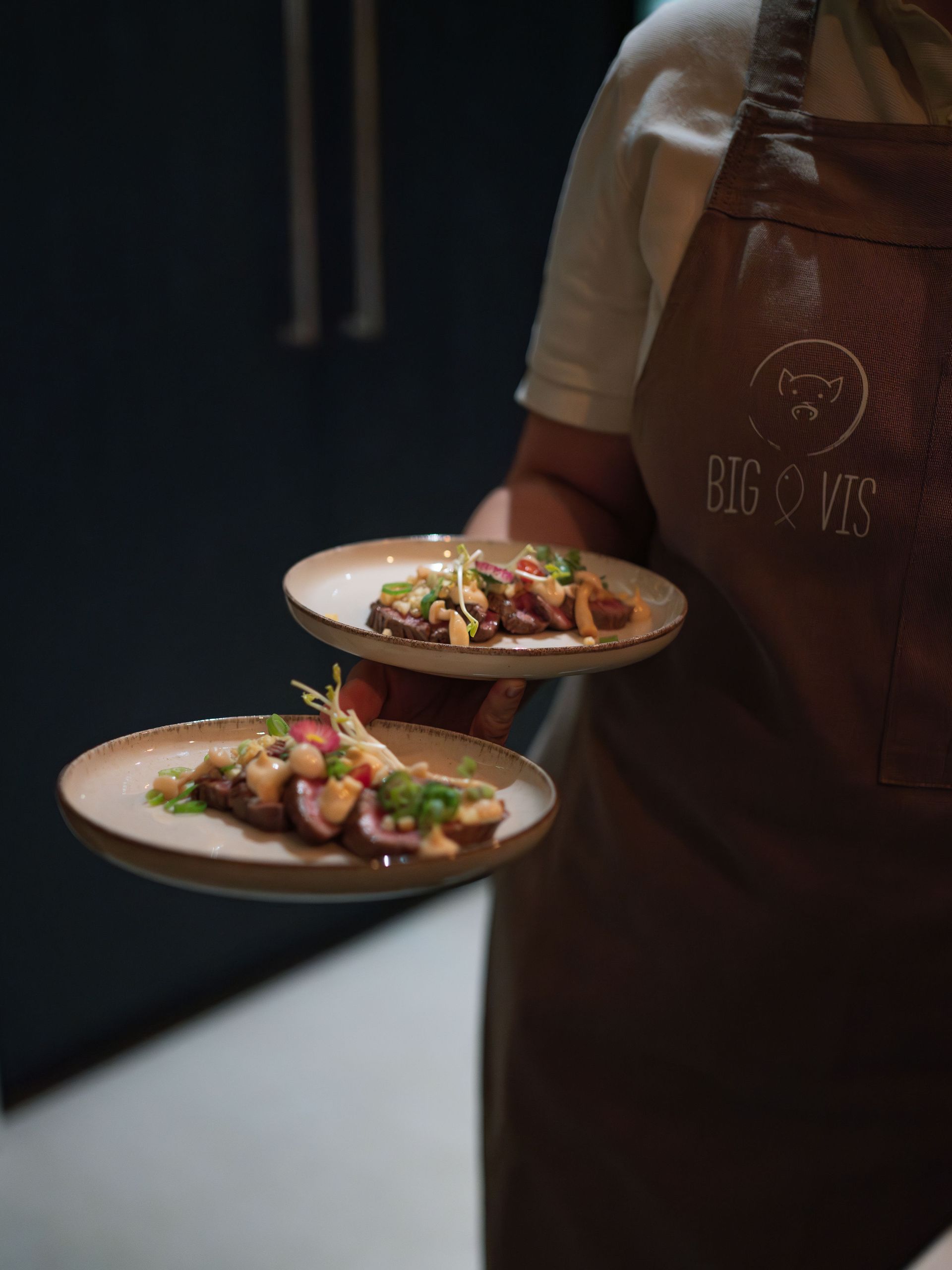 Een persoon met een bruin schort houdt twee borden vast met kleine porties gebakken vlees, gegarneerd met verschillende toppings.