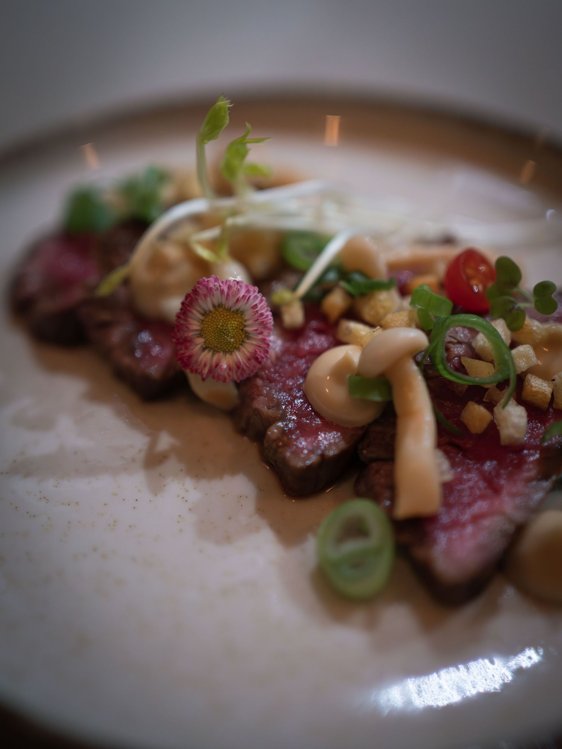 Een close-up van dun gesneden, medium-rare biefstuk, gegarneerd met champignons, andere ingrediënten en een klein bloemetje, op een wit bord.