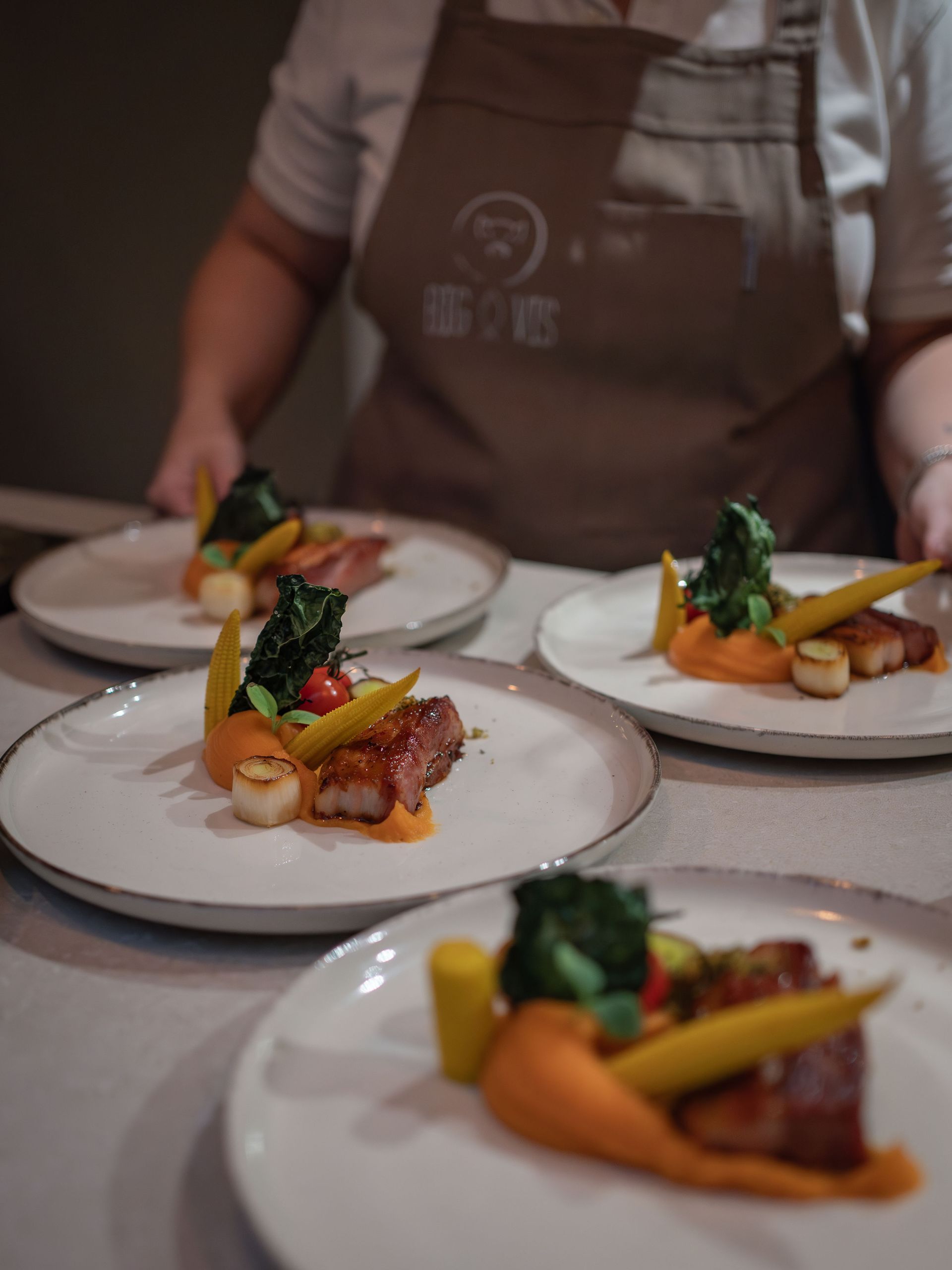 Iemand met een beige schort zet borden neer met daarop gebakken vlees, sint-jakobsschelpen, groentepuree en garnering.