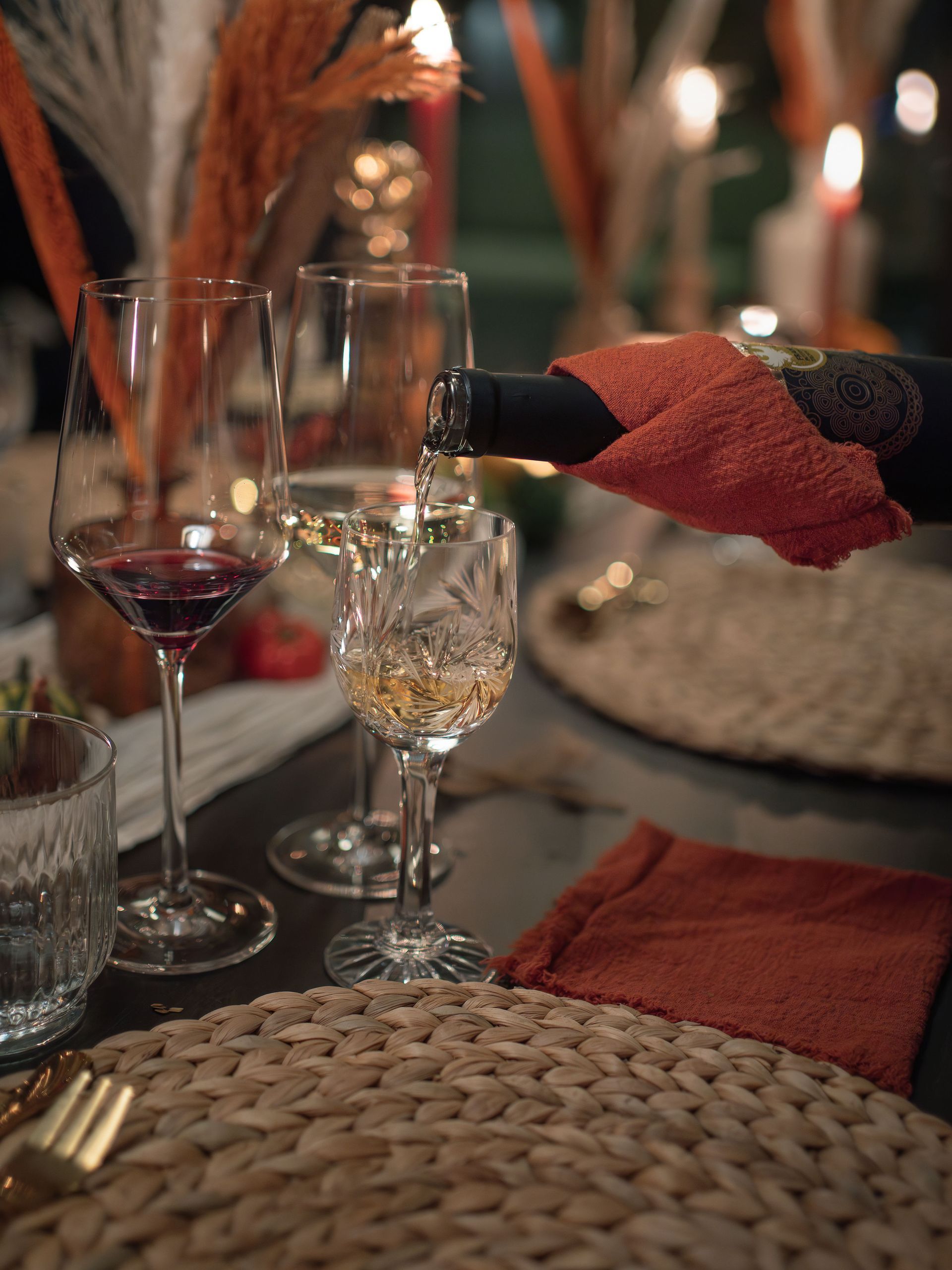 Een hand schenkt witte wijn uit een donkere fles in een kristallen glas aan een tafel bij kaarslicht, gedekt met geweven placemats.