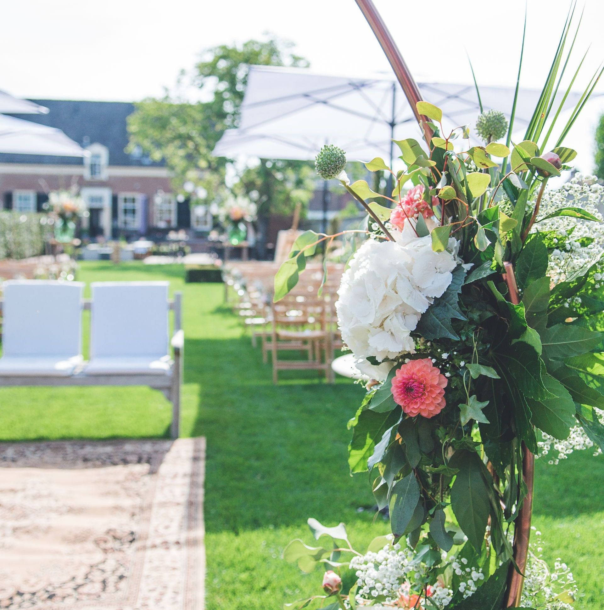 Een bloemstuk staat op de voorgrond van een buitenbruiloft op een groen gazon met stoelen en parasols.