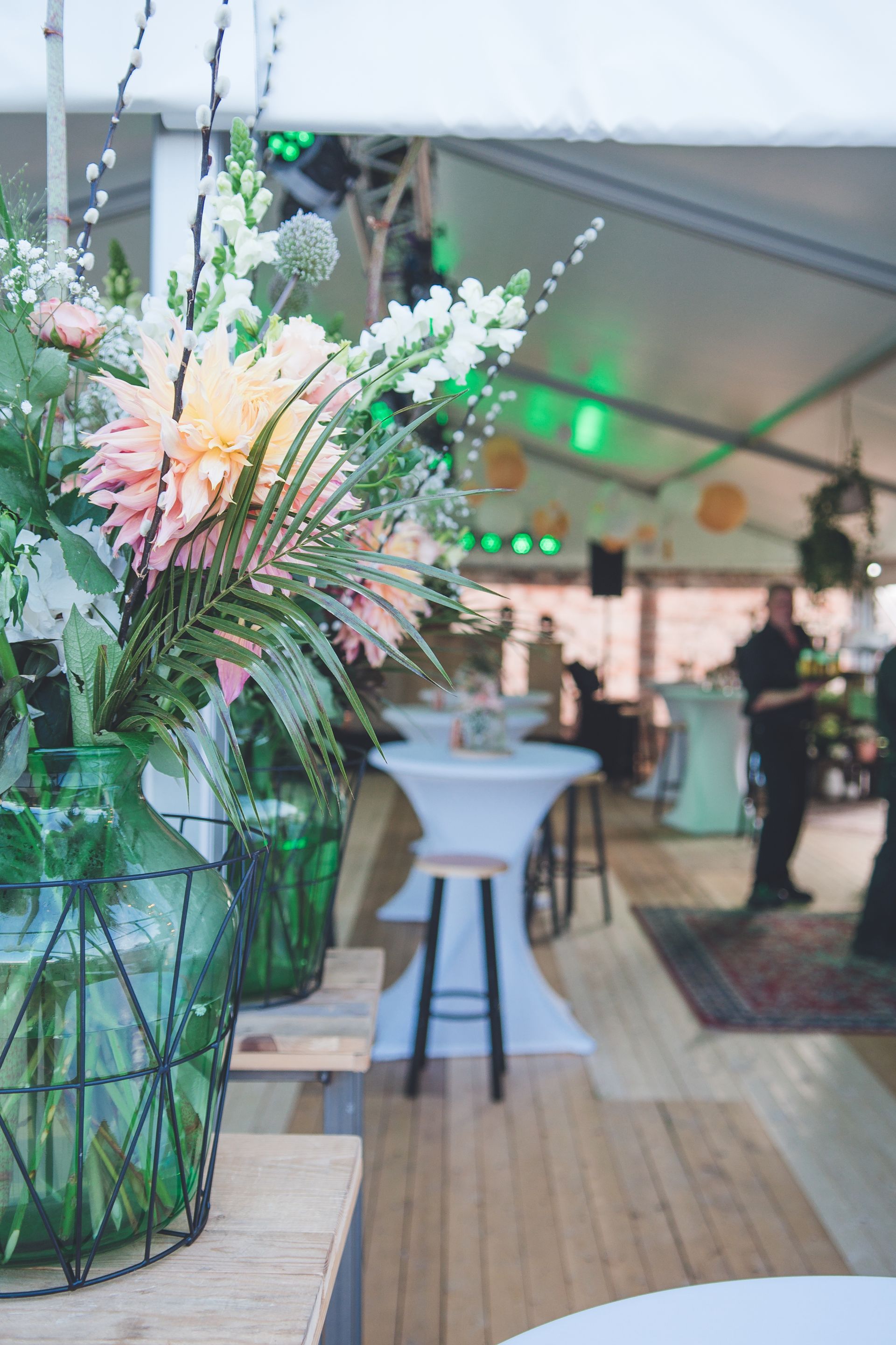 Een groot bloemstuk in een groene glazen vaas staat op een houten tafel in een tentvormige evenementenruimte met hoge cocktailtafels.