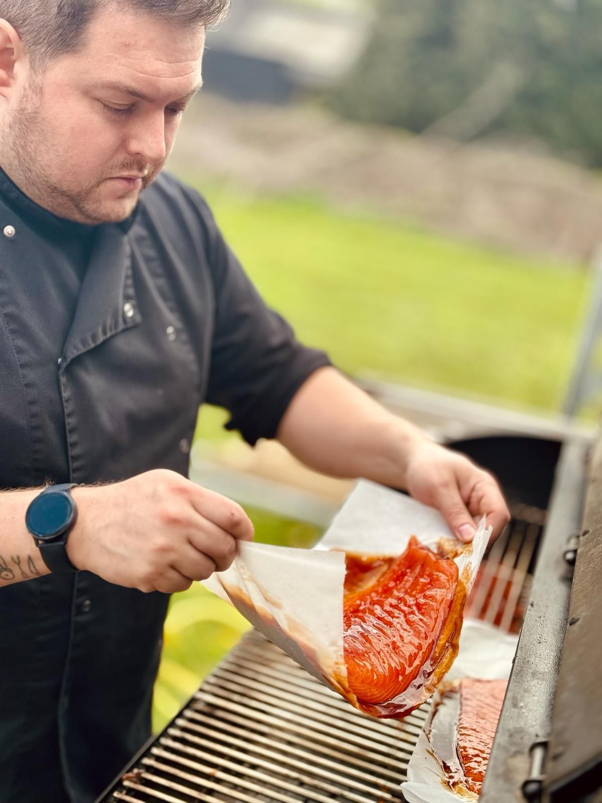 Een chef-kok in een zwart uniform tilt een stuk geglazuurde, gebakken zalm van bakpapier boven een buitengrill.