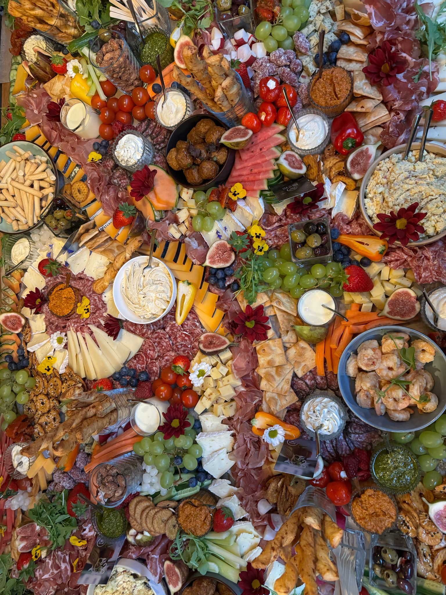 Een grote, rijkelijk gevulde charcuterieplank met vleeswaren, kazen, druiven, fruit, noten, dipsauzen, crackers en eetbare bloemen.