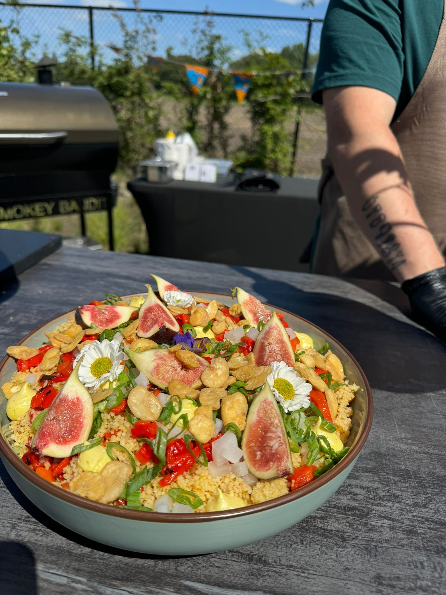 Een kom gele graansalade, gegarneerd met plakjes verse vijgen, witte bloemen en knapperige stukjes, staat buiten op een tafel.