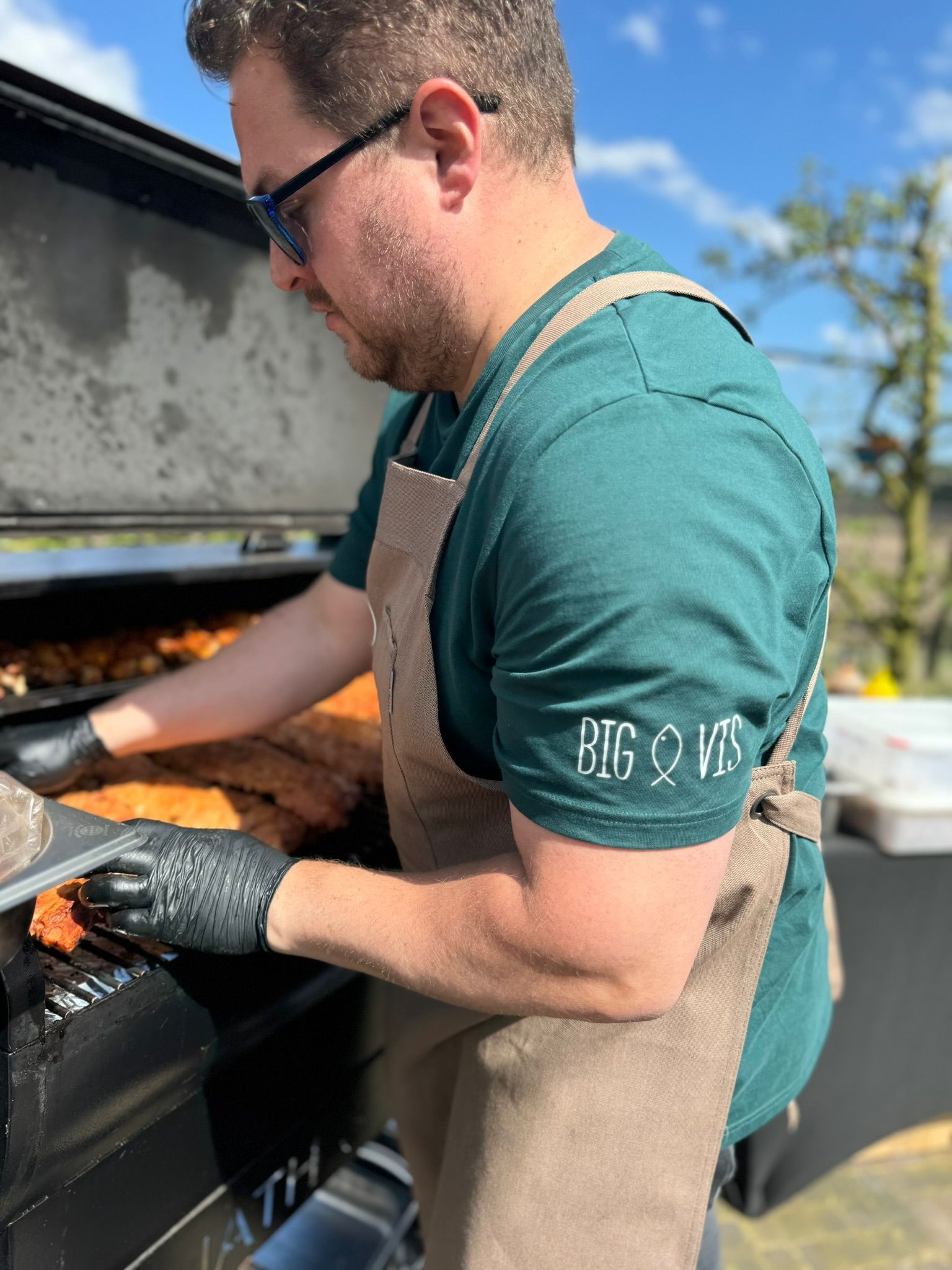 Een persoon, gekleed in een groen T-shirt en een beige schort, gebruikt zwarte handschoenen om vlees te grillen op een buitenbarbecue.