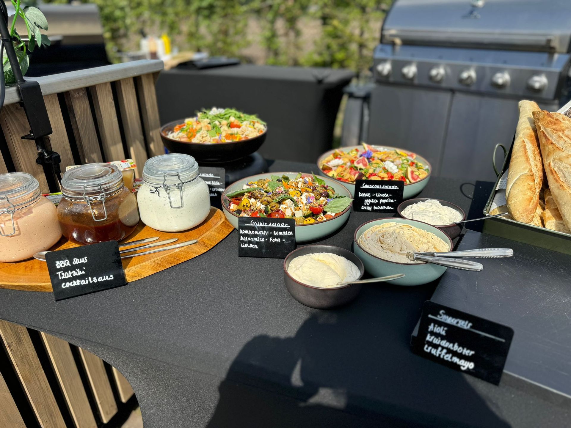 Een buffettafel met schalen salade, dipsauzen en knapperig brood buiten, vlakbij een barbecue.