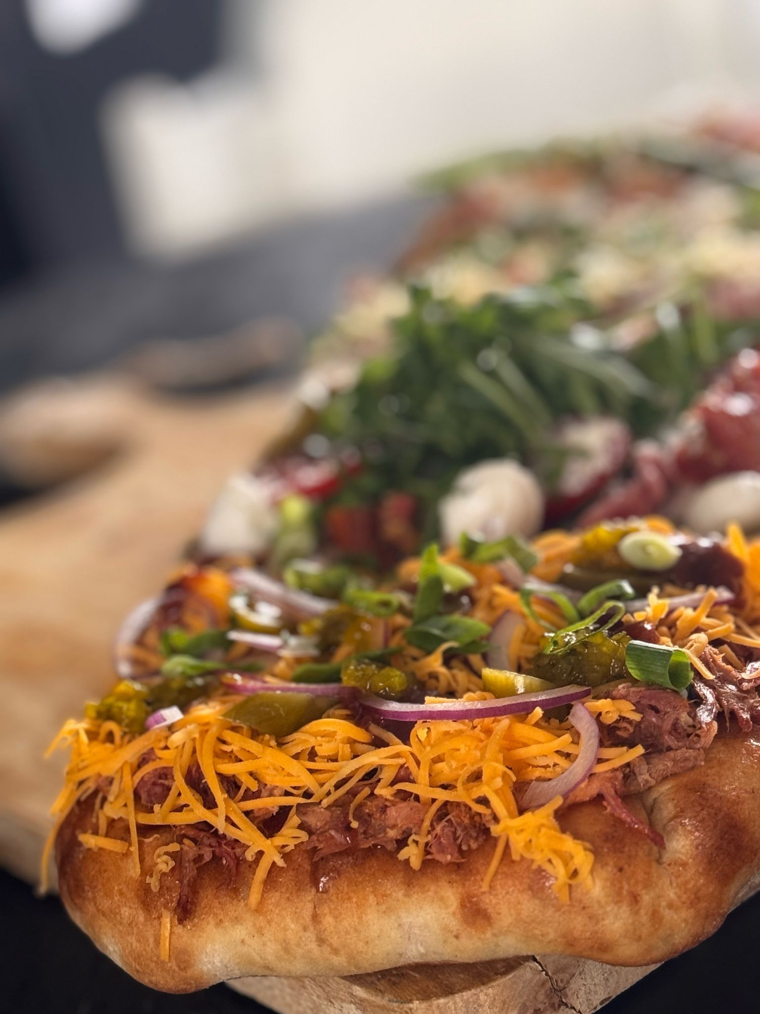 Twee platbroodpizza's op een houten plank, de ene belegd met cheddar, rode ui en jalapeño's, de andere met groenten en vlees.