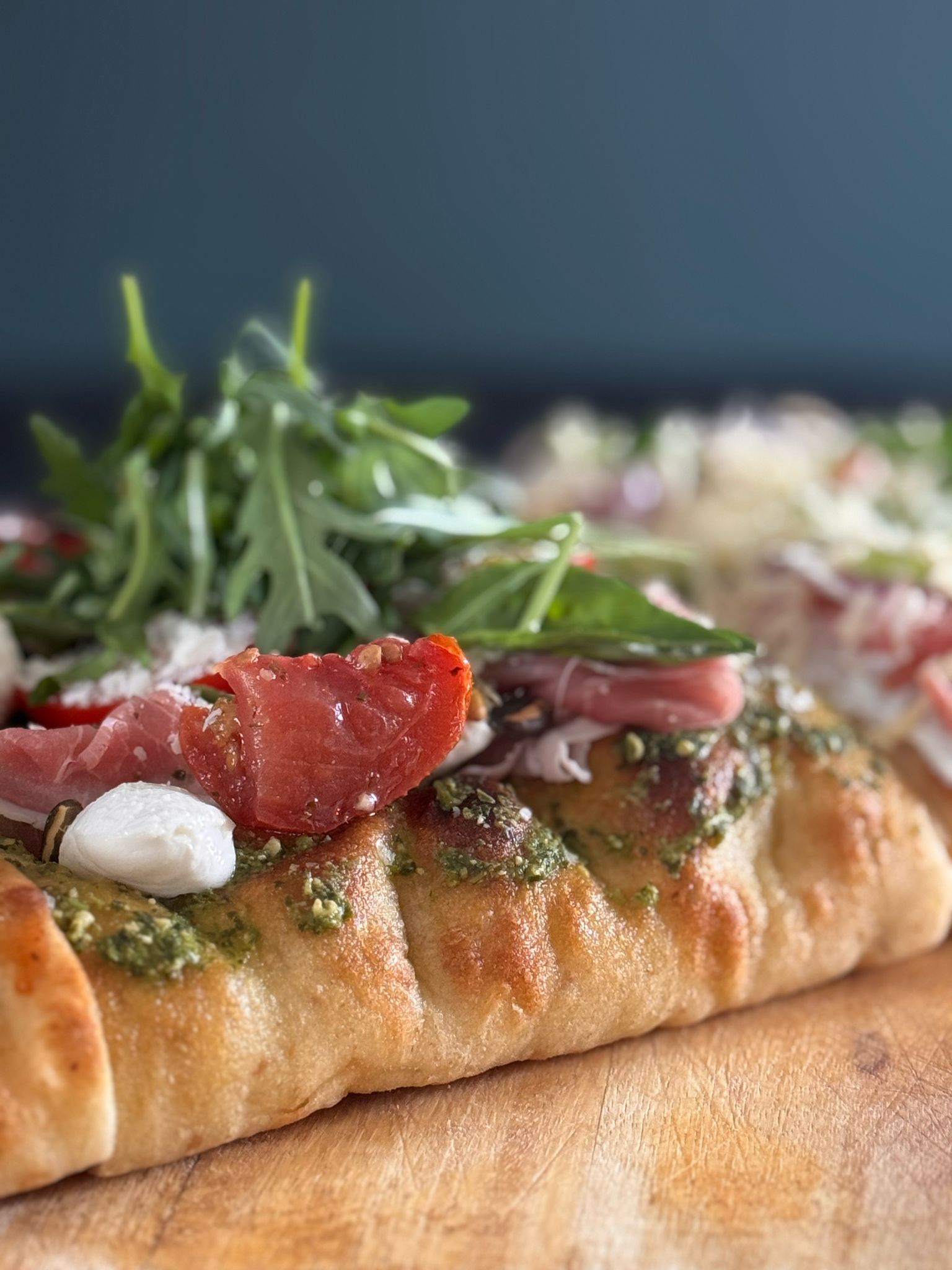 Close-up van een platbrood belegd met pesto, prosciutto, verse mozzarella en rucola op een houten plank.